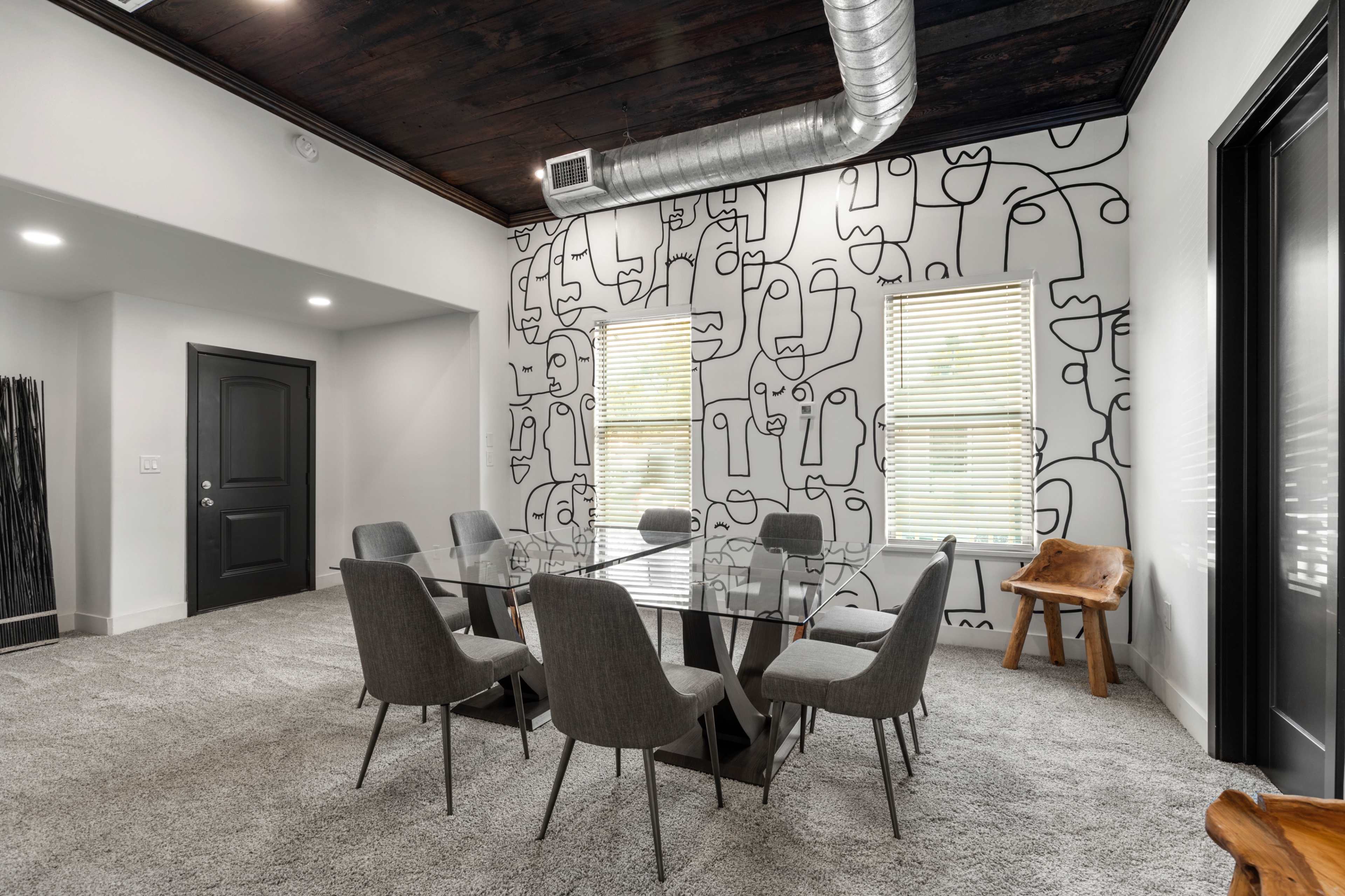 A dining area features a glass table surrounded by six chairs, with a wall adorned by a minimalist line drawing design and a wooden accent piece in the corner.