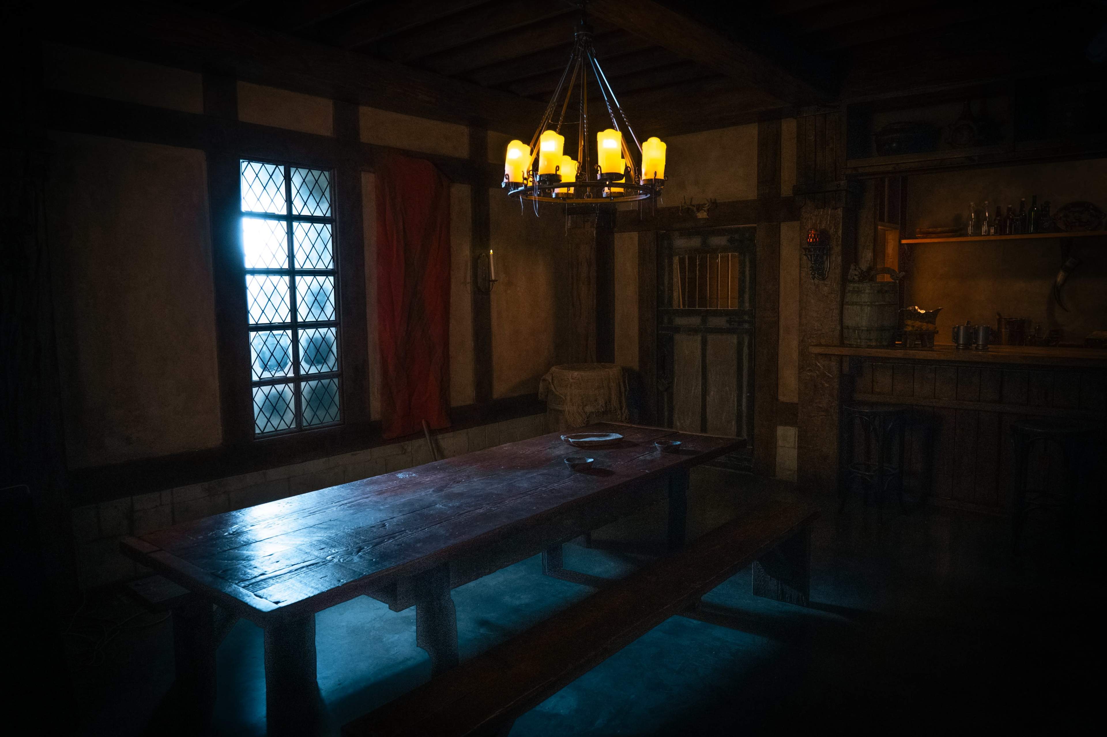 The image shows a dimly lit medieval-style room featuring a large wooden table, a chandelier with candles, and a window with a red curtain.
