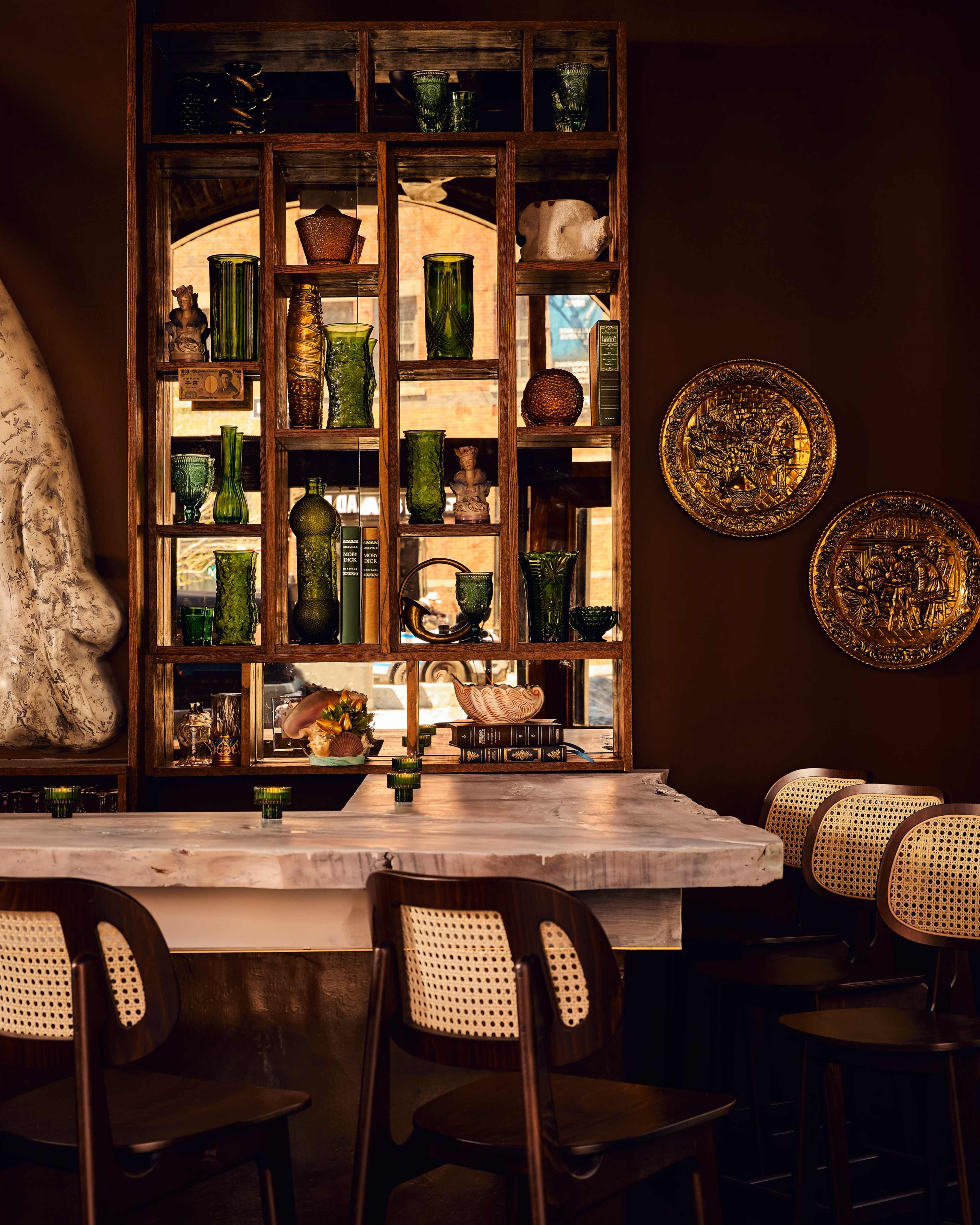 The image shows a bar area with a marble countertop and wooden cabinetry displaying decorative items and green glass vases against a darkly colored wall.
