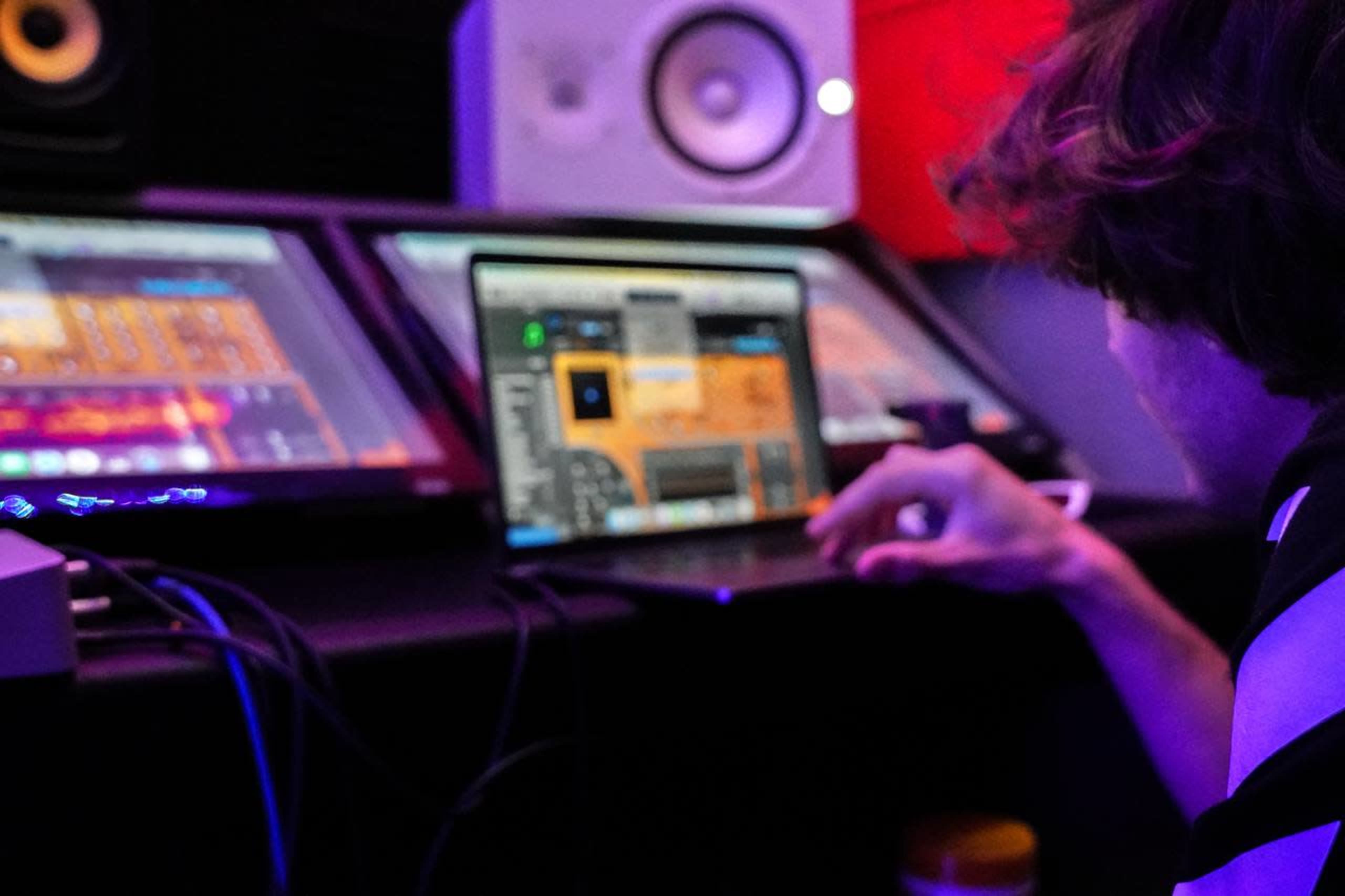A person is working on a laptop surrounded by audio equipment in a music production studio.