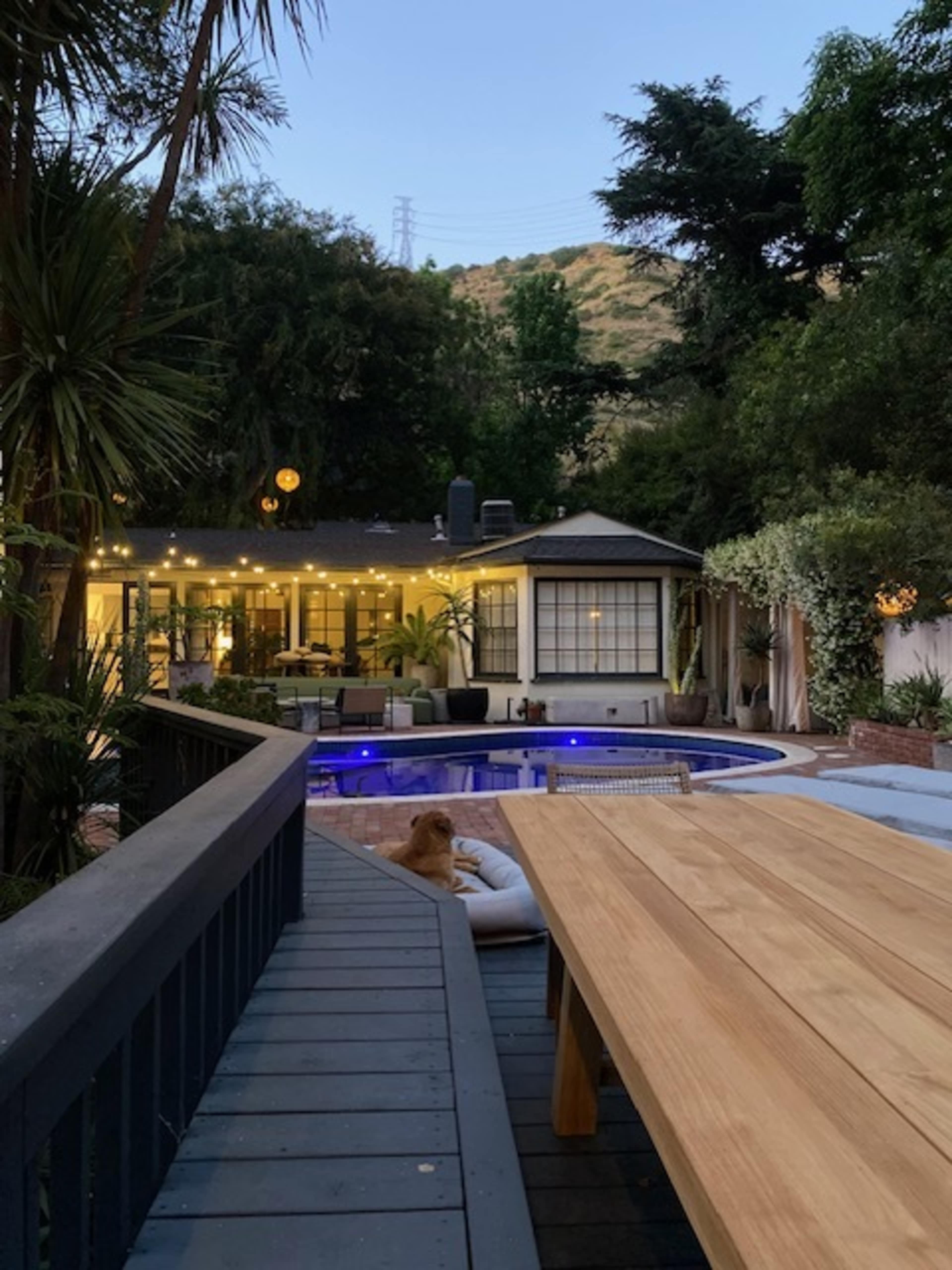 A wooden table is positioned on a deck overlooking a pool and a well-lit backyard with a house in the background.