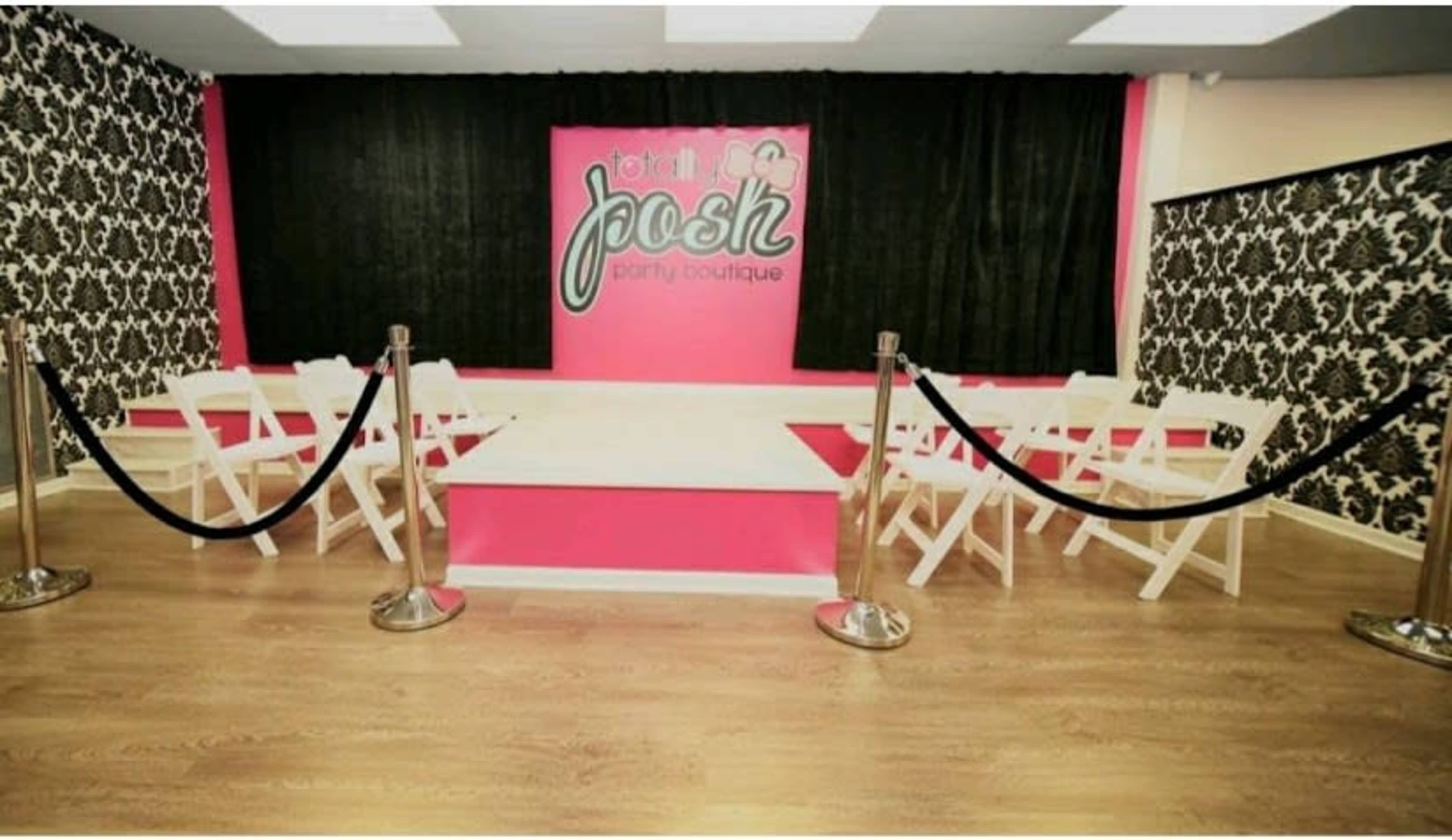 A party boutique interior featuring a runway-style stage, white folding chairs arranged in front, and a vibrant pink and black color scheme on the walls.