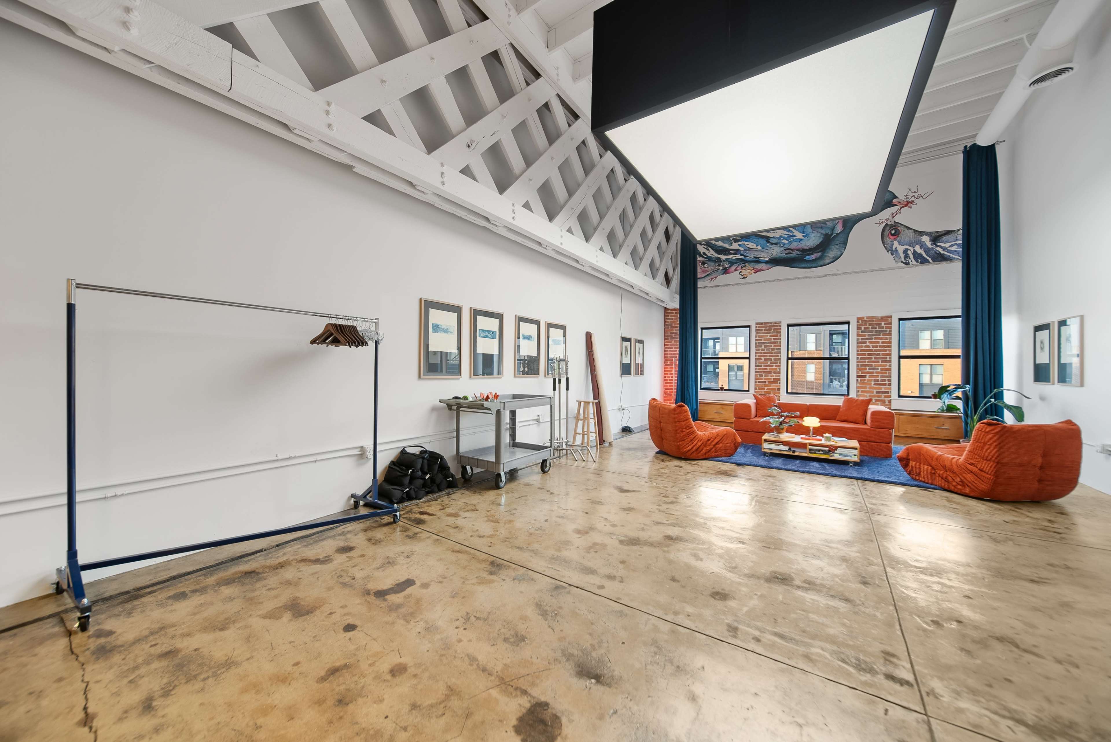 The image shows a spacious interior with concrete flooring, featuring an orange sofa arrangement, a coffee table, and wall-mounted artwork, along with a coat rack and a cart in the corner.