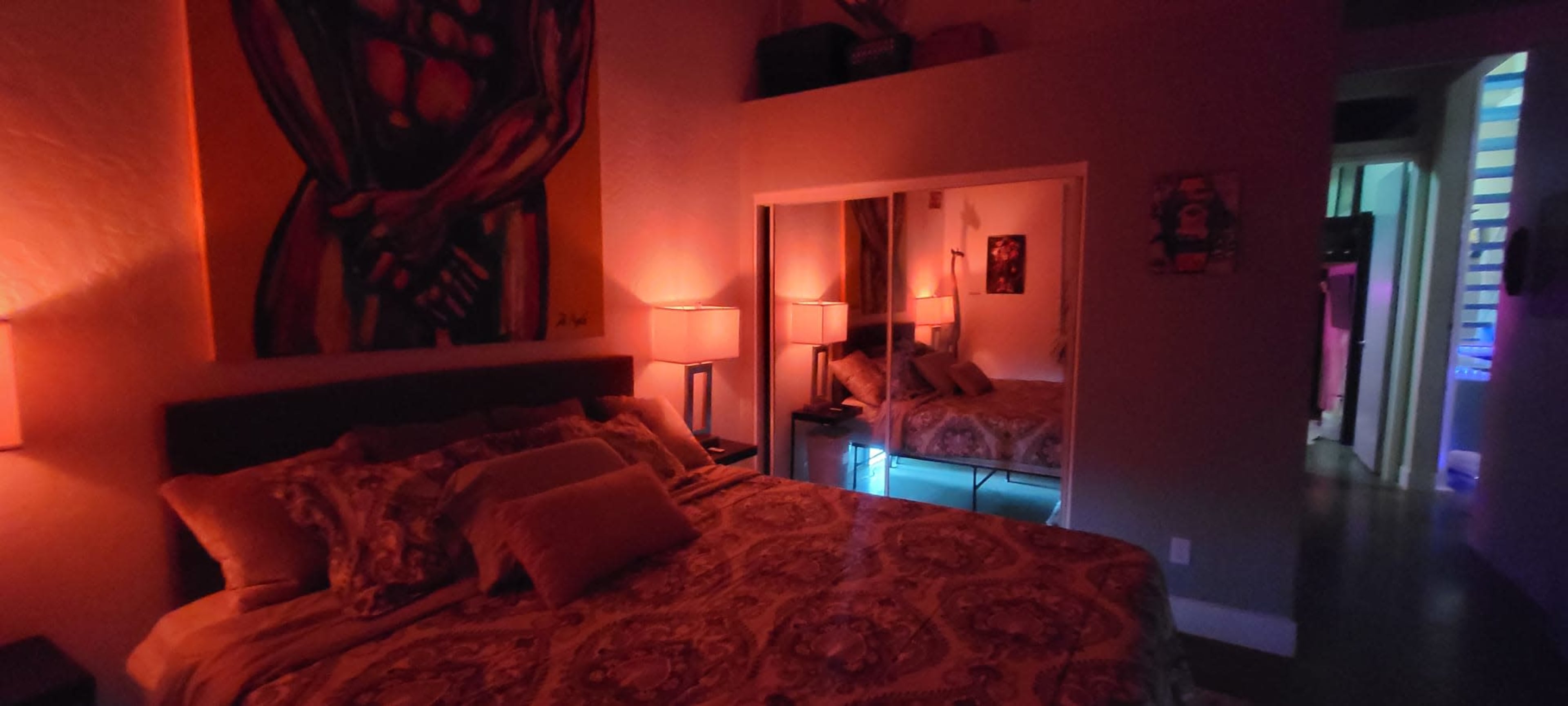 A dimly-lit bedroom with a large bed, decorative pillows, and artwork on the walls, featuring a mirror that reflects the room's subtle lighting.