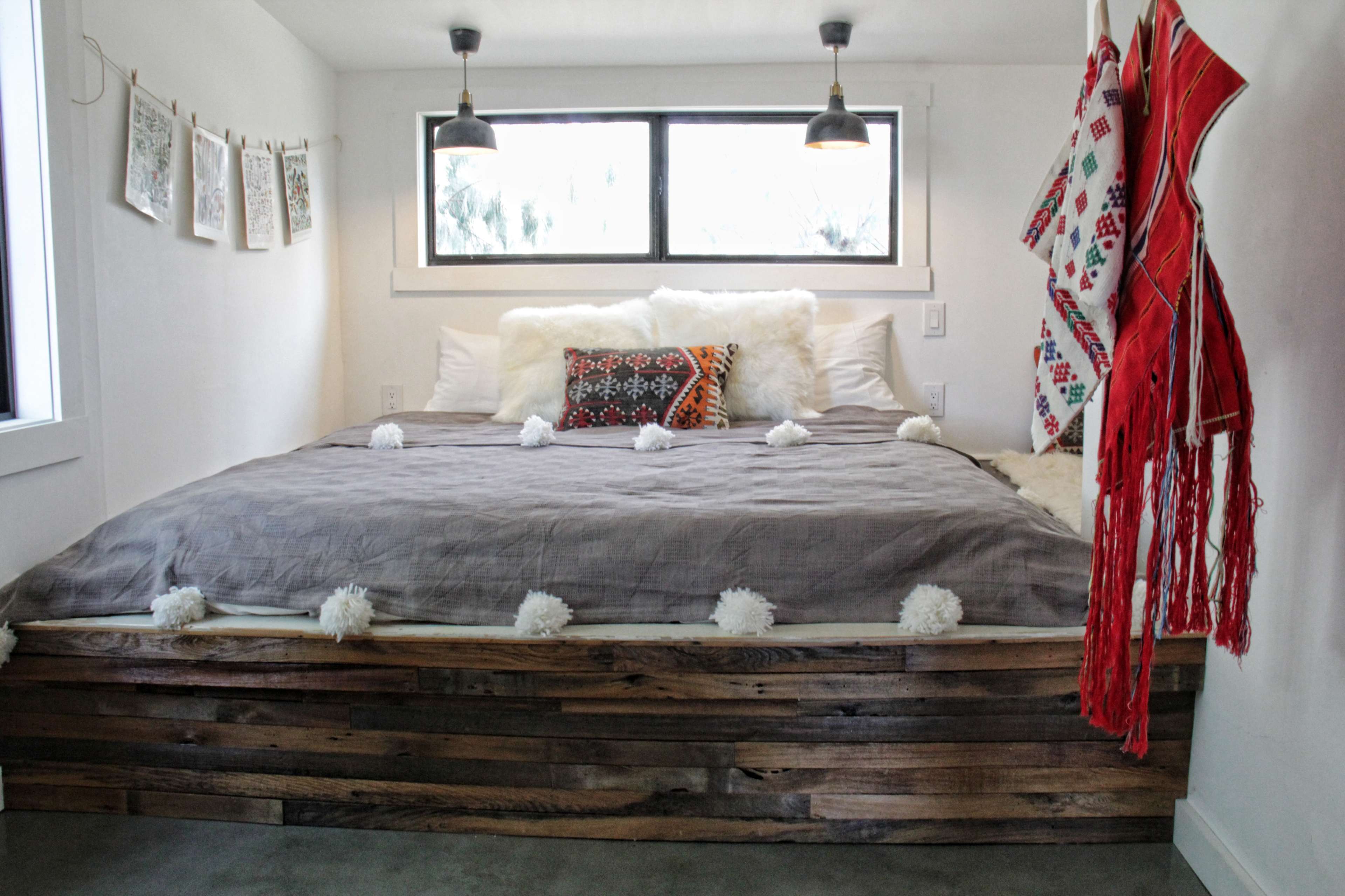 A cozy bedroom with a raised wooden bed, adorned with a gray blanket and decorative pillows, alongside white fur throws and wall art.