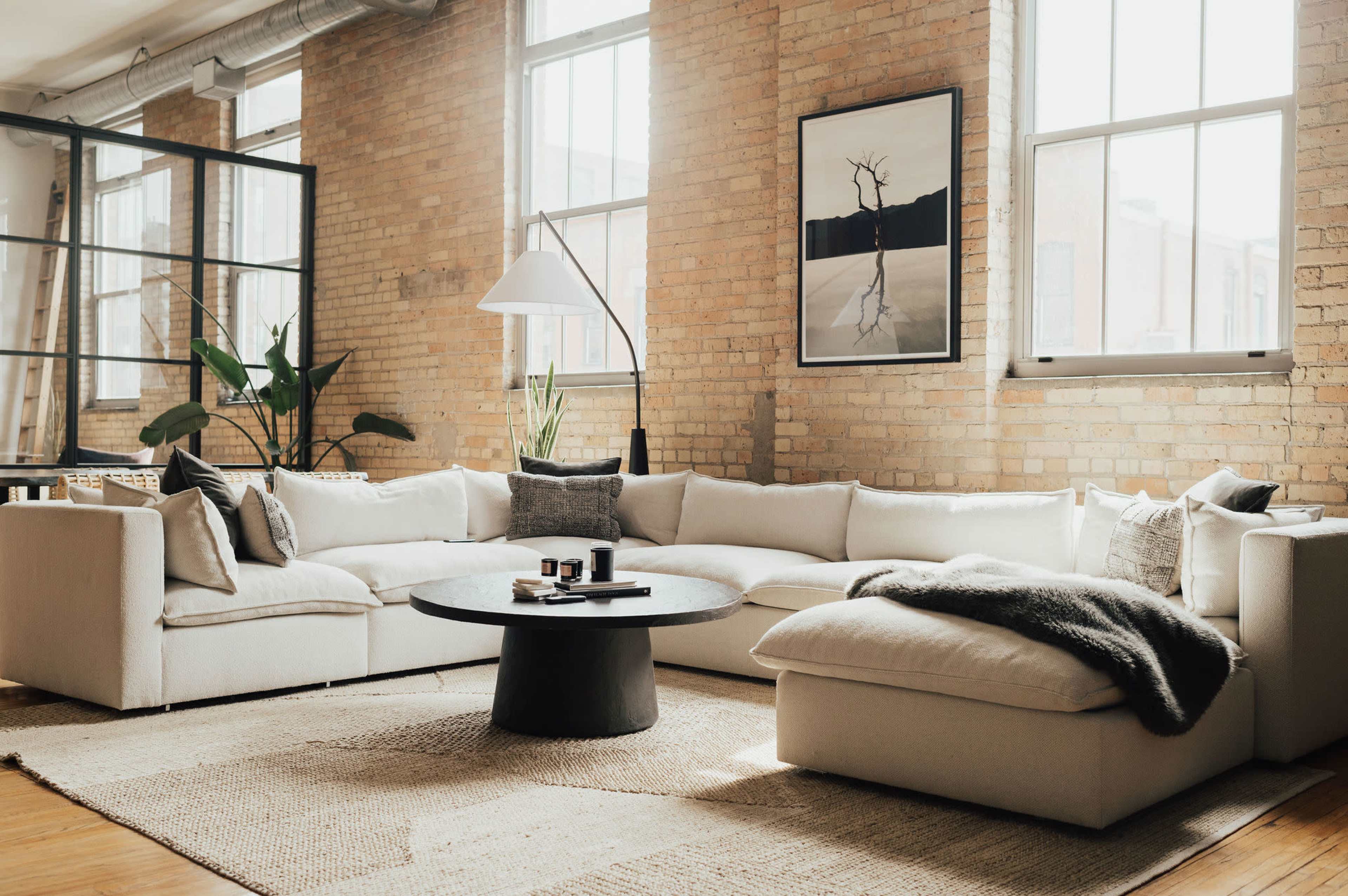 A bright, modern living room with a large white sectional sofa, a round black coffee table, and large windows allowing natural light to fill the space.