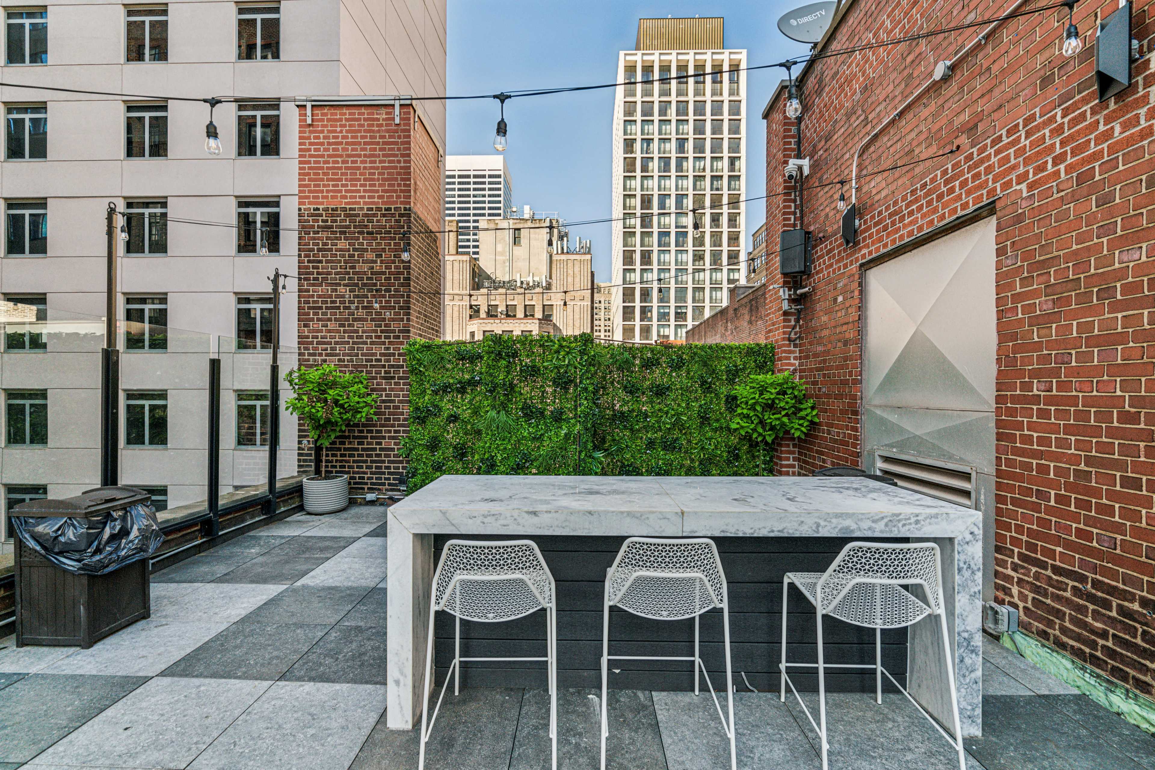 A rooftop terrace features a large marble table with three white wire chairs, surrounded by greenery and tall buildings in the background.