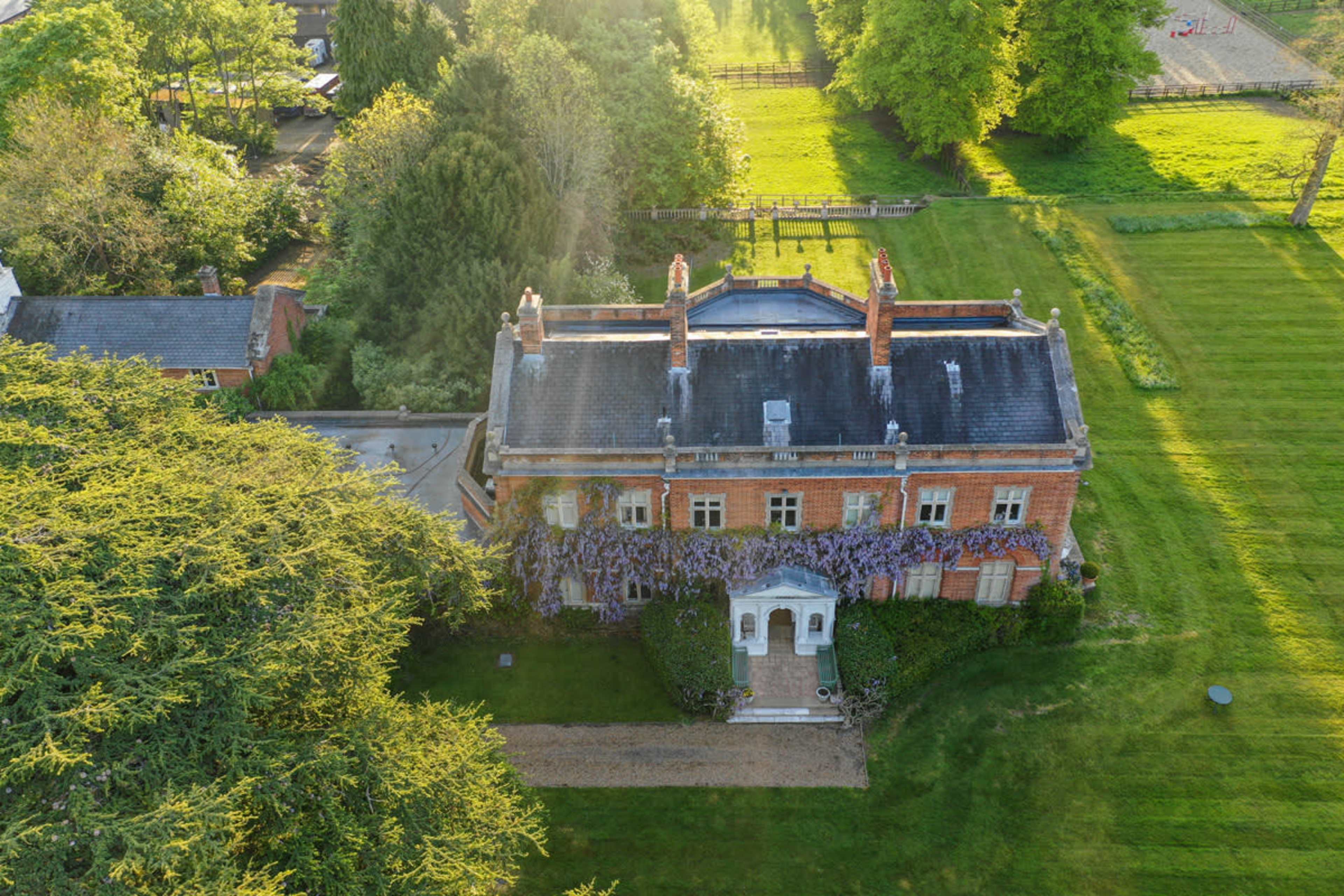 An aerial view of a large, brick house with a wide front staircase, surrounded by green lawns and trees.