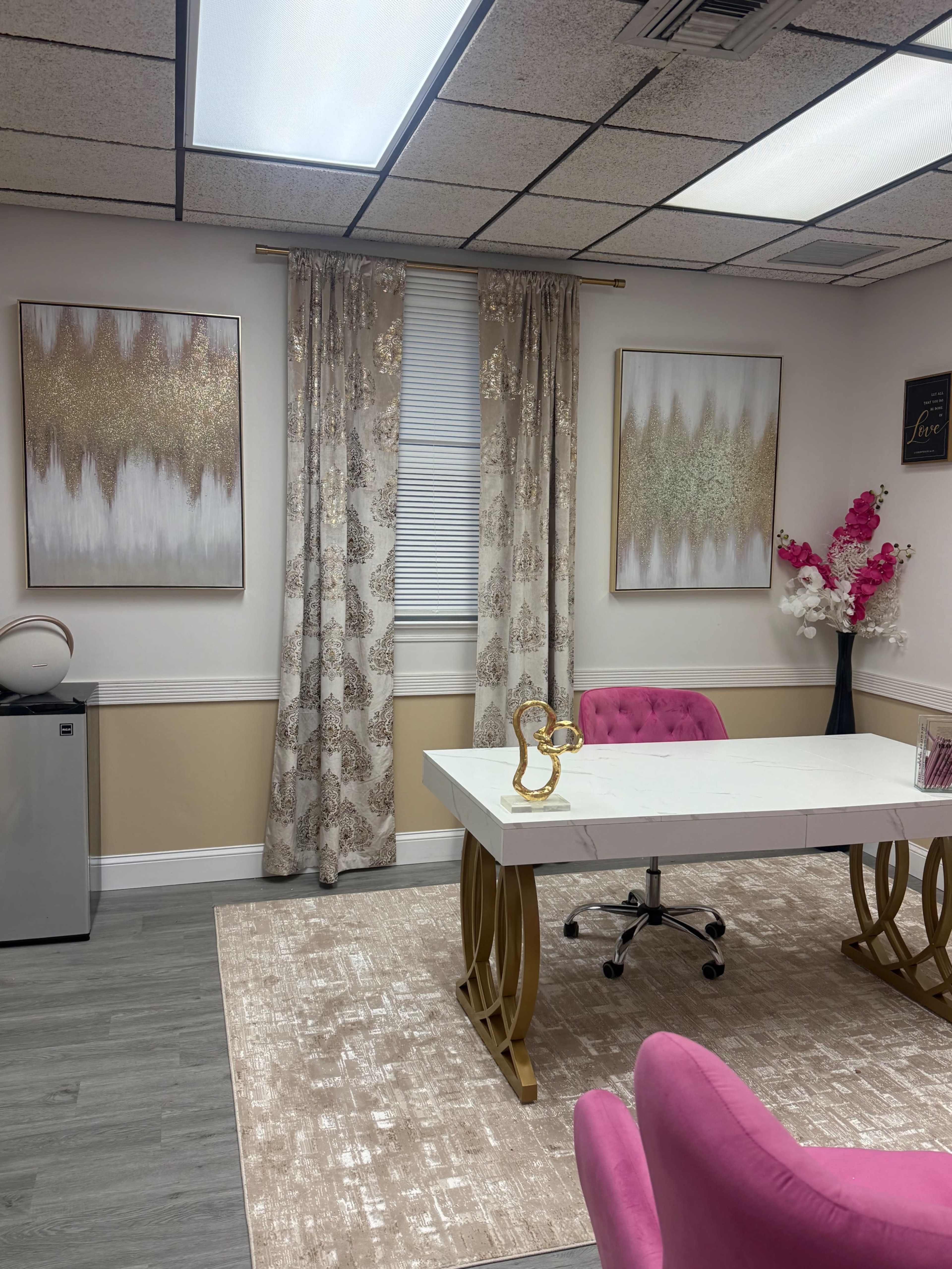 The image shows a modern office space featuring a white desk with a gold base, pink accent chairs, gold and white wall art, and curtains adorned with a floral pattern.