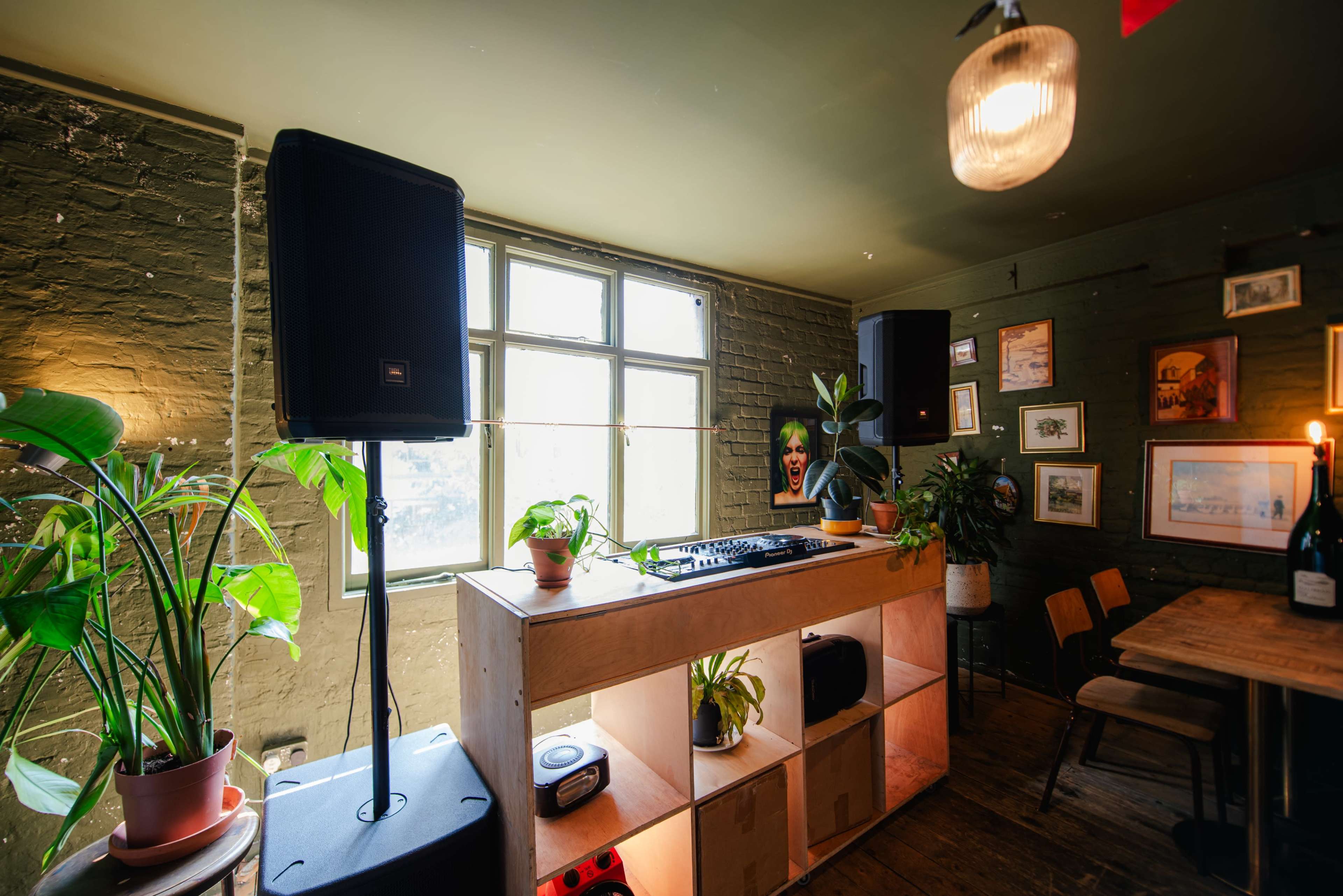 The image shows a cozy interior of a bar featuring a DJ setup on a wooden stand surrounded by potted plants and soft lighting.