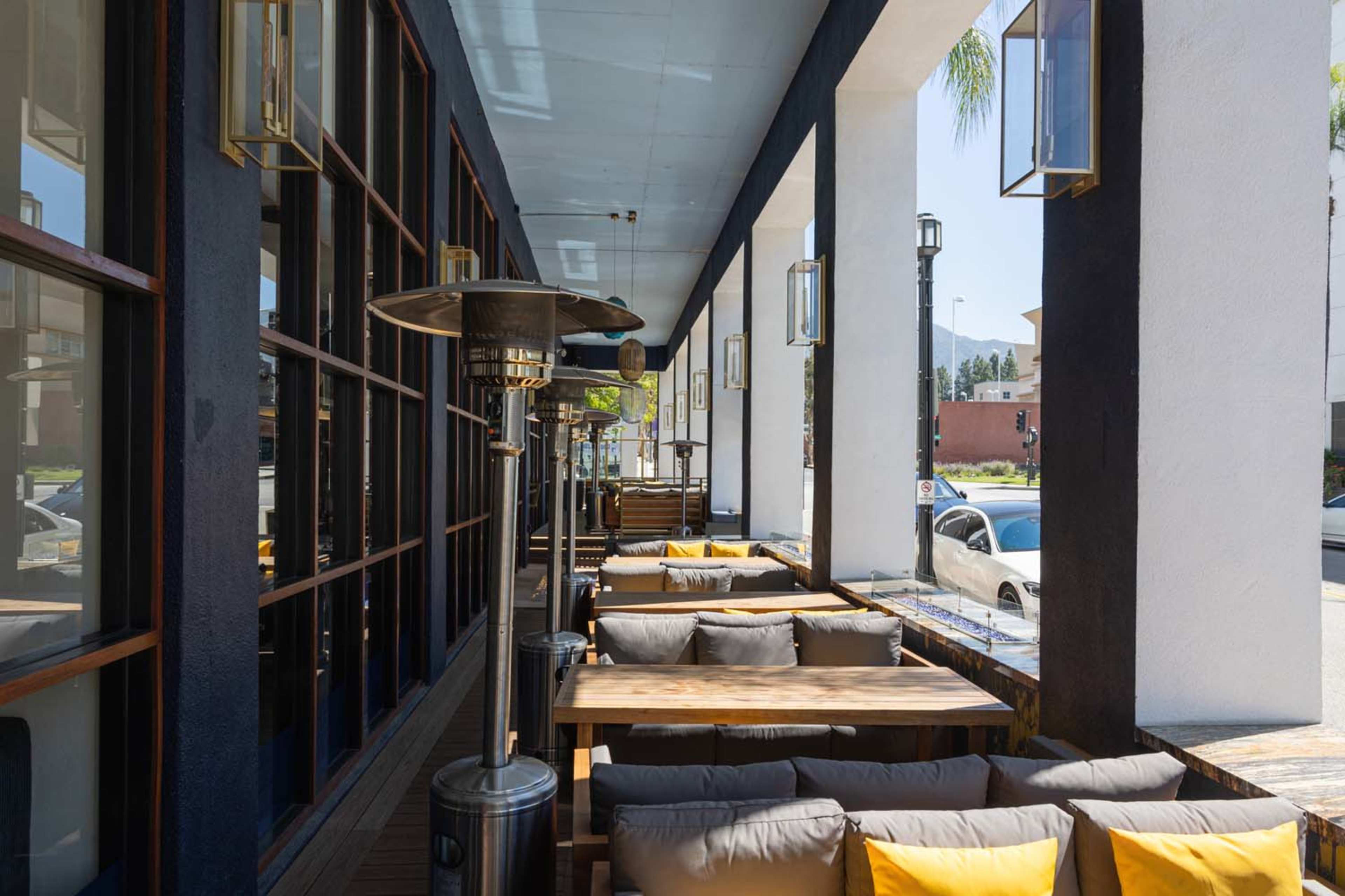 The image shows an outdoor dining area with wooden tables and cushioned seating, flanked by large windows and patio heaters under a clear blue sky.