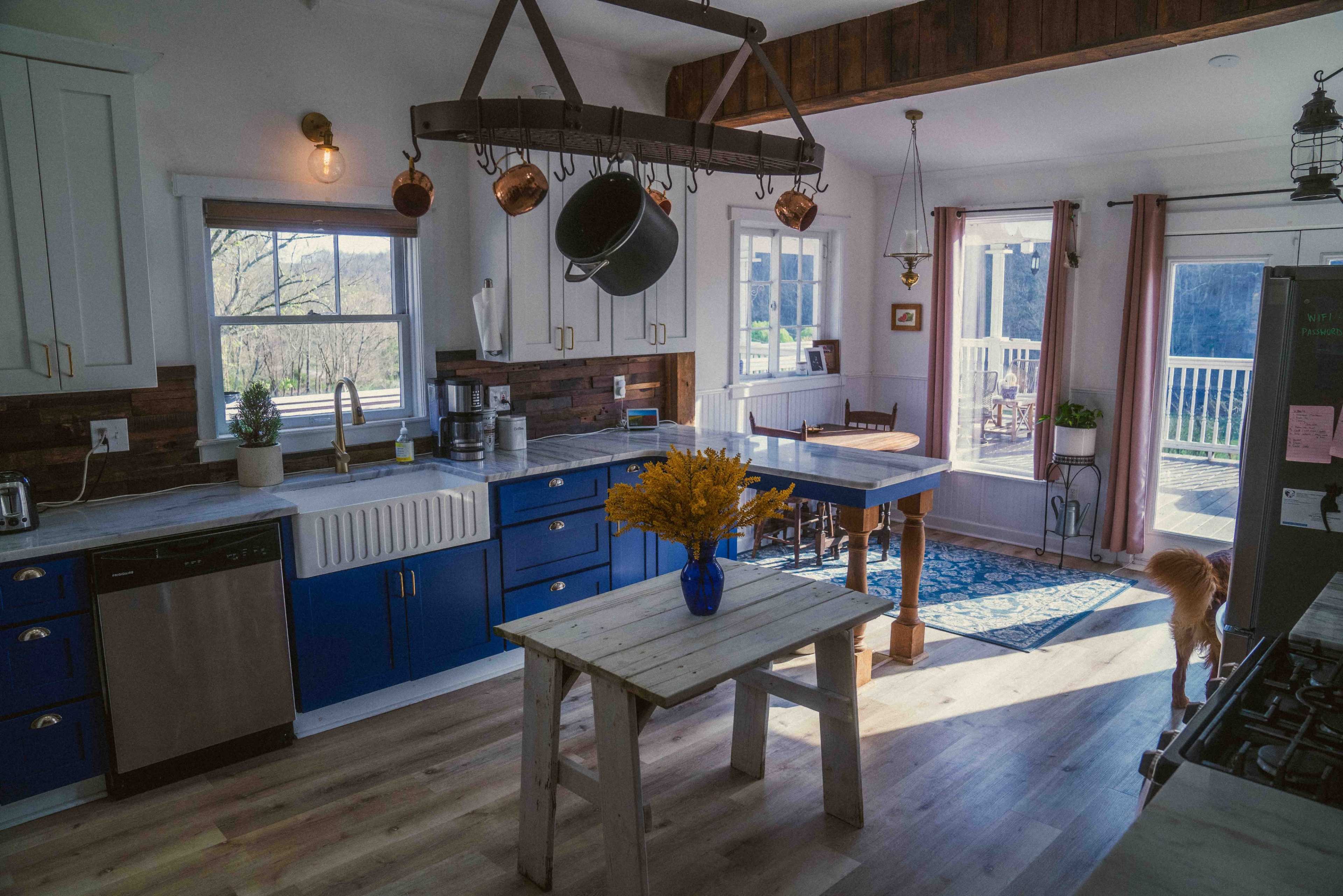 The image shows a bright, modern kitchen featuring a blue and white color scheme, with a central table, hanging pots, and access to a deck through glass doors.