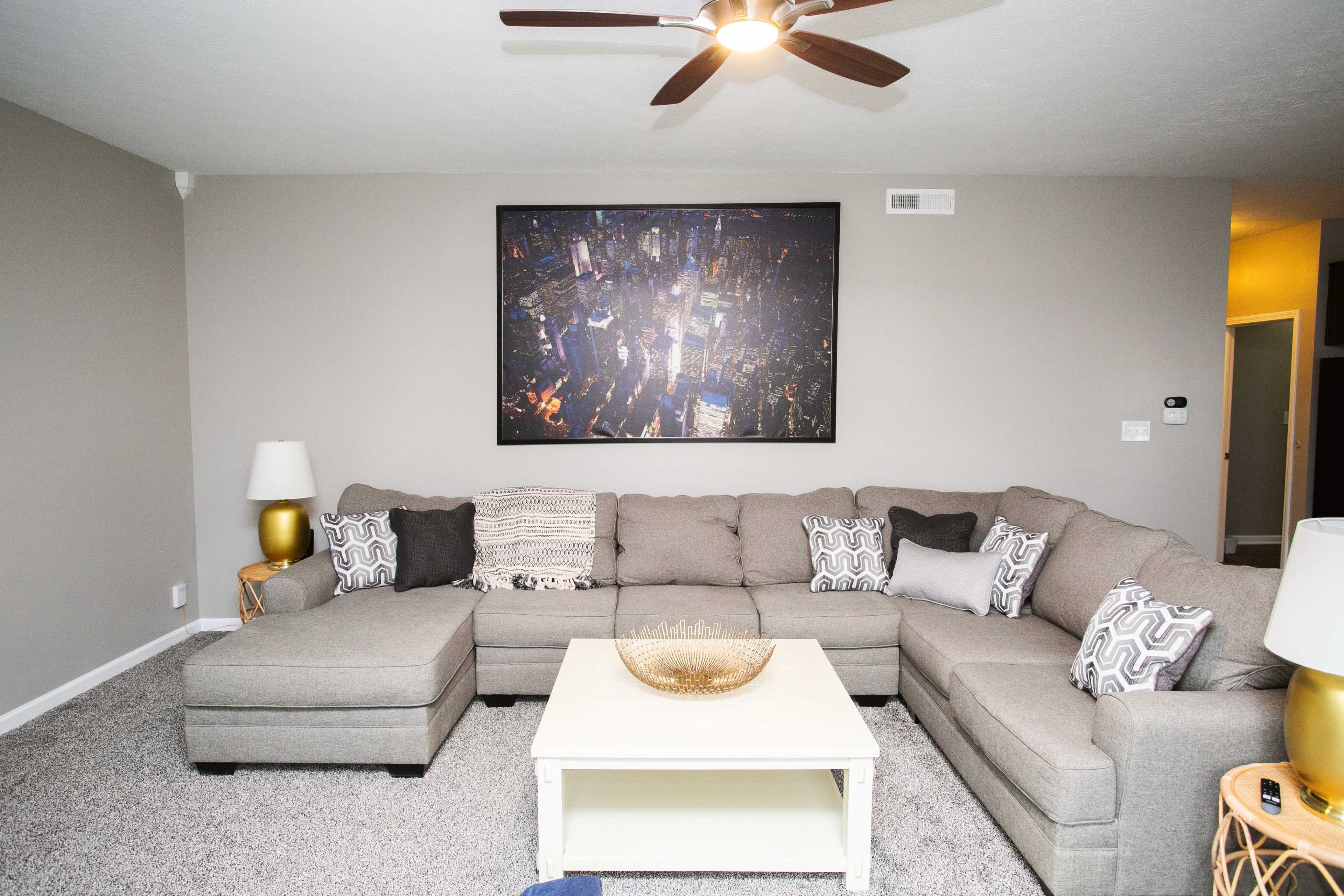 A modern living room features a large sectional sofa, a coffee table, and a wall-mounted cityscape photo.