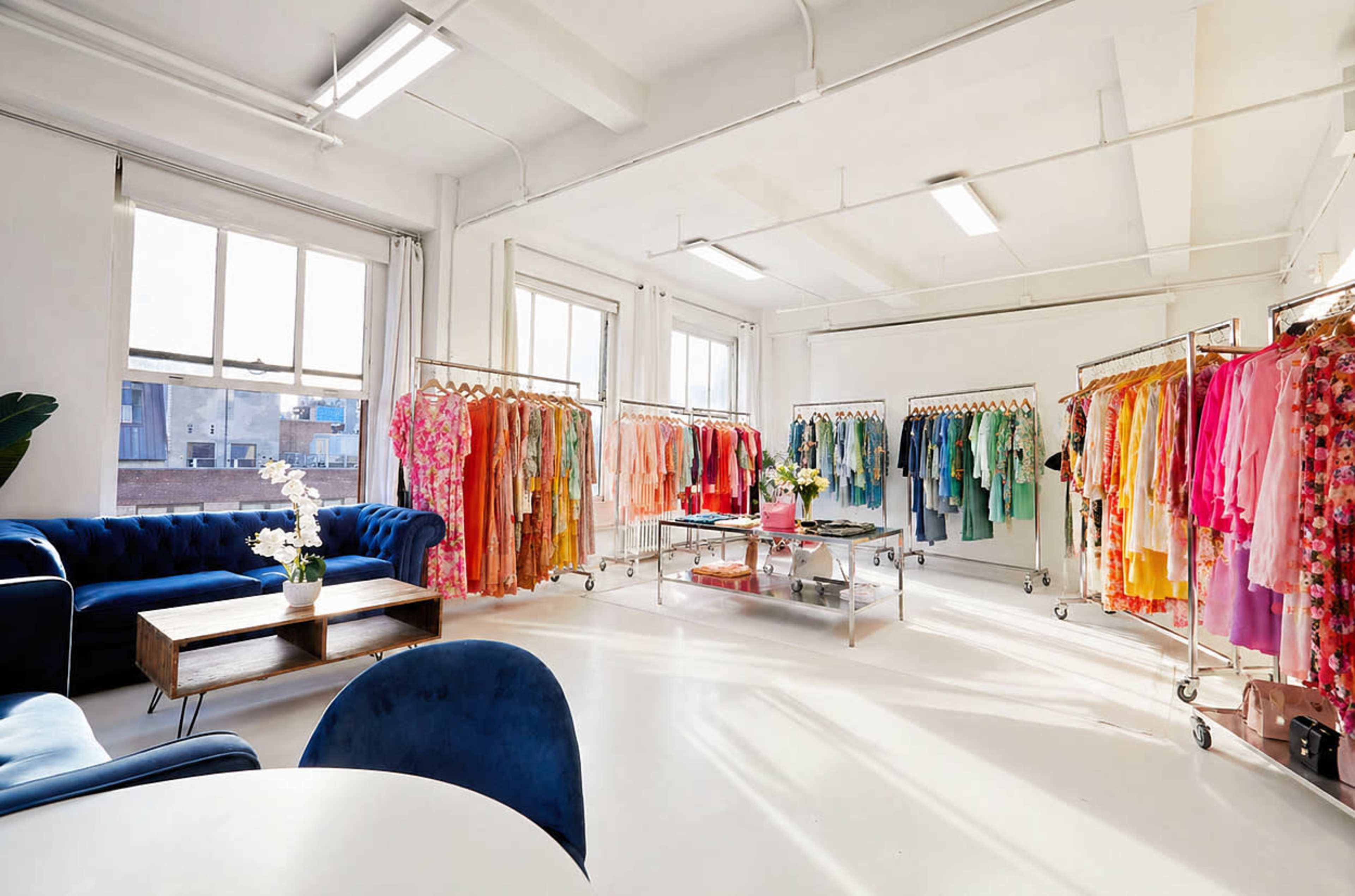 The image shows a bright, spacious room with clothing racks filled with colorful garments and a seating area featuring a blue sofa and coffee table.