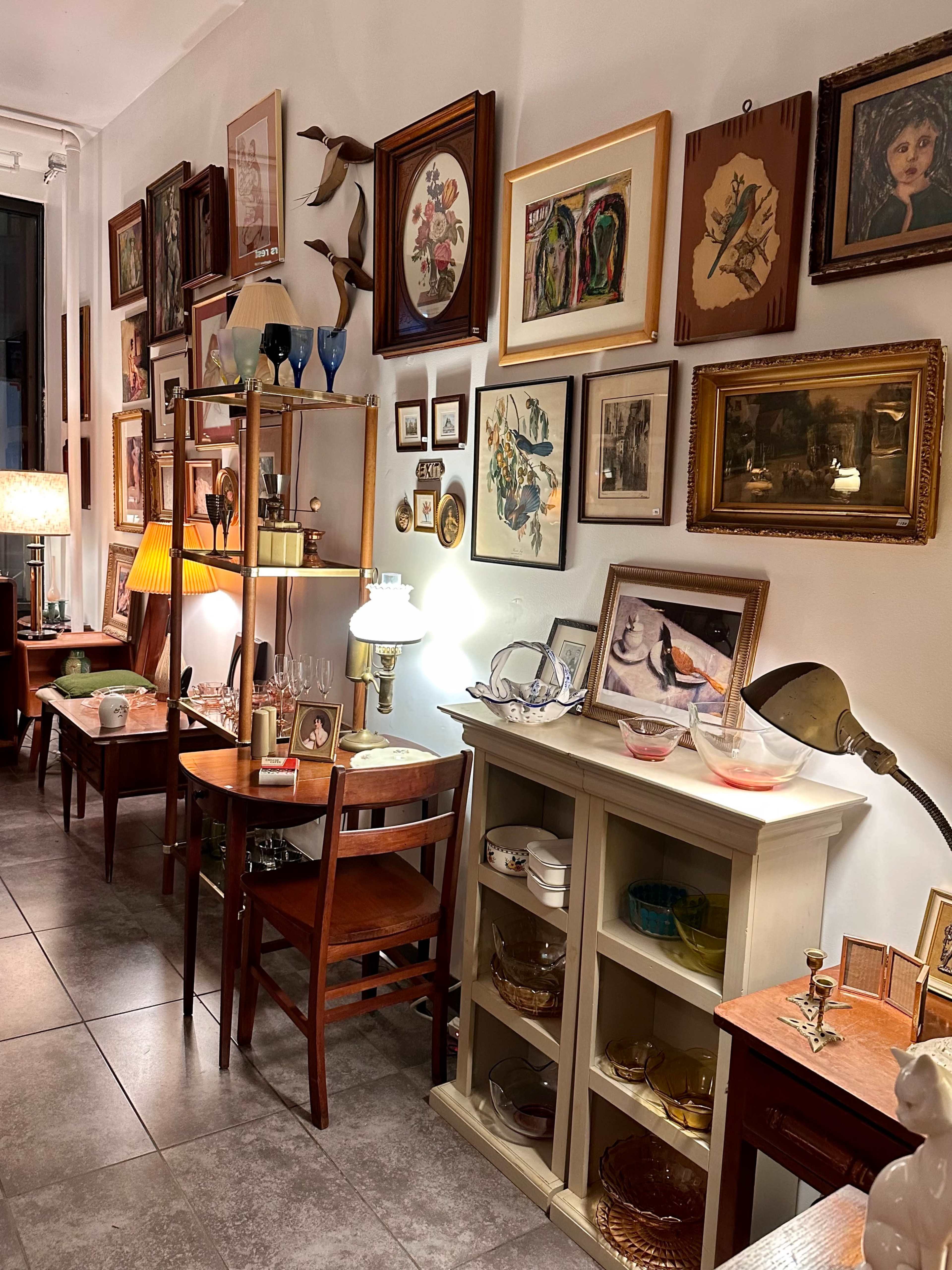 The image shows a cozy room filled with various pieces of vintage furniture, framed artwork, and decorative items arranged on walls and shelves.