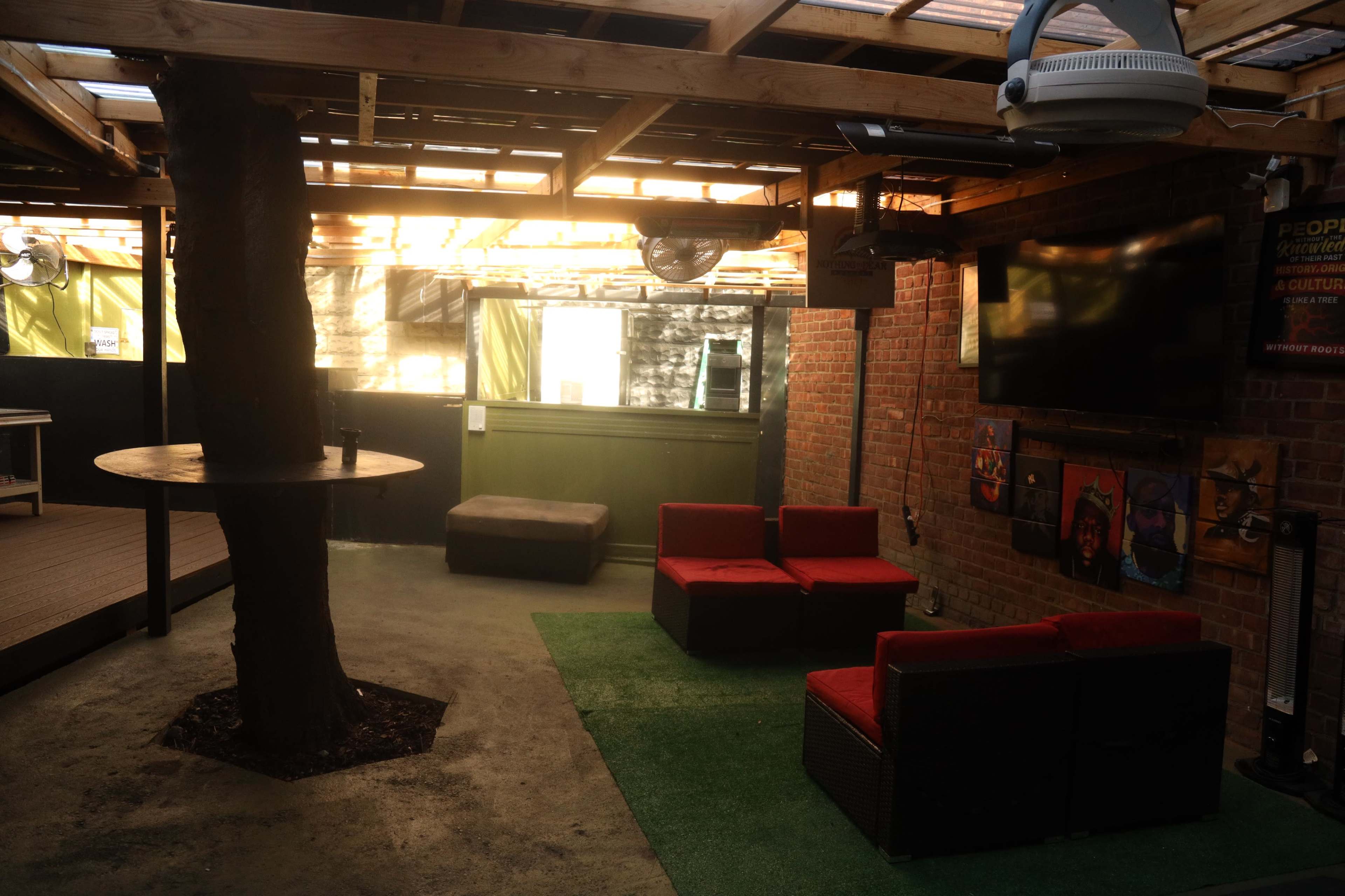 An indoor lounge area with a large tree in the center, red seating, a TV mounted on the wall, and an outdoor view visible through the open space.