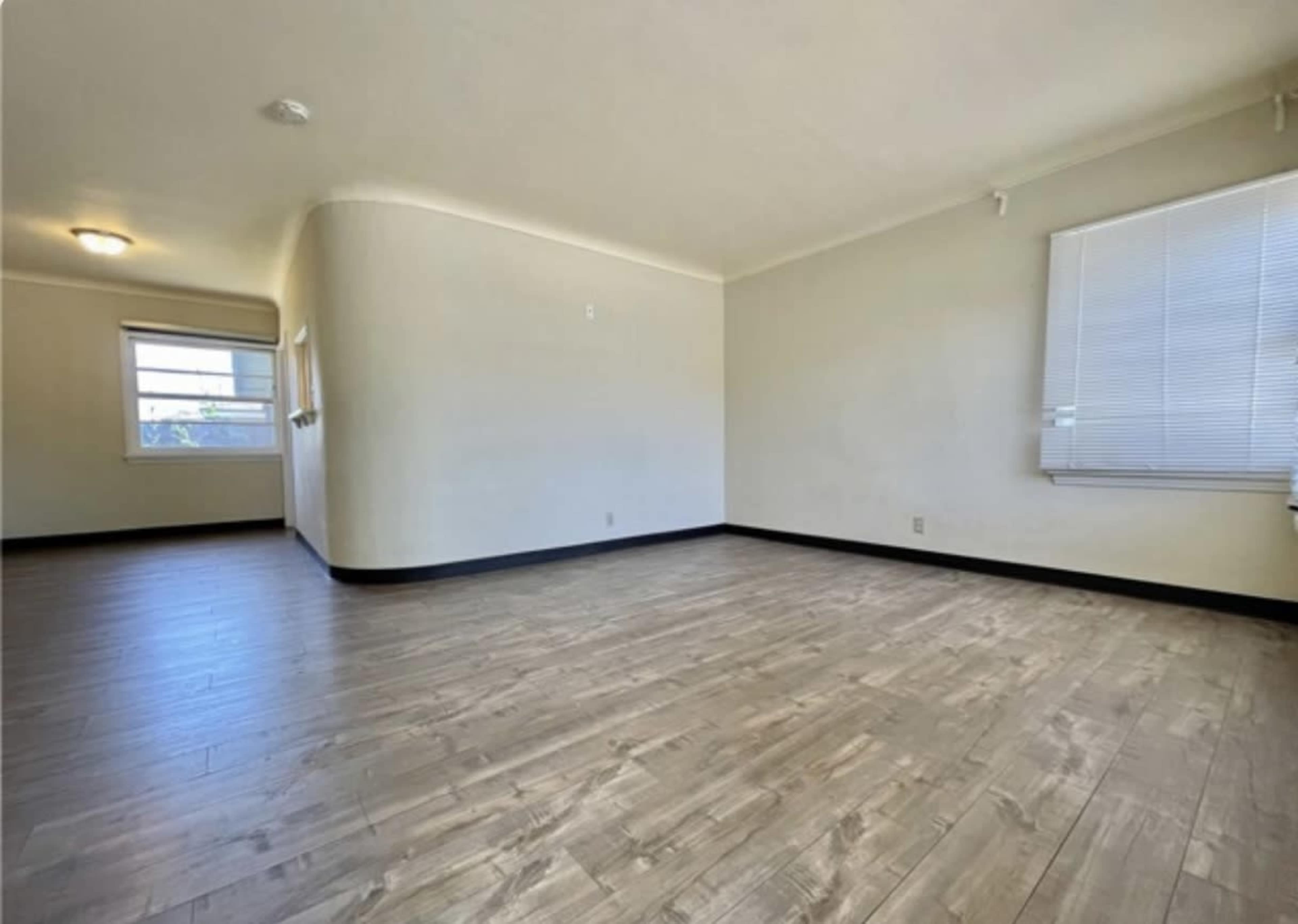 The image shows a spacious, empty room with hardwood-like flooring, large windows, and plain walls.