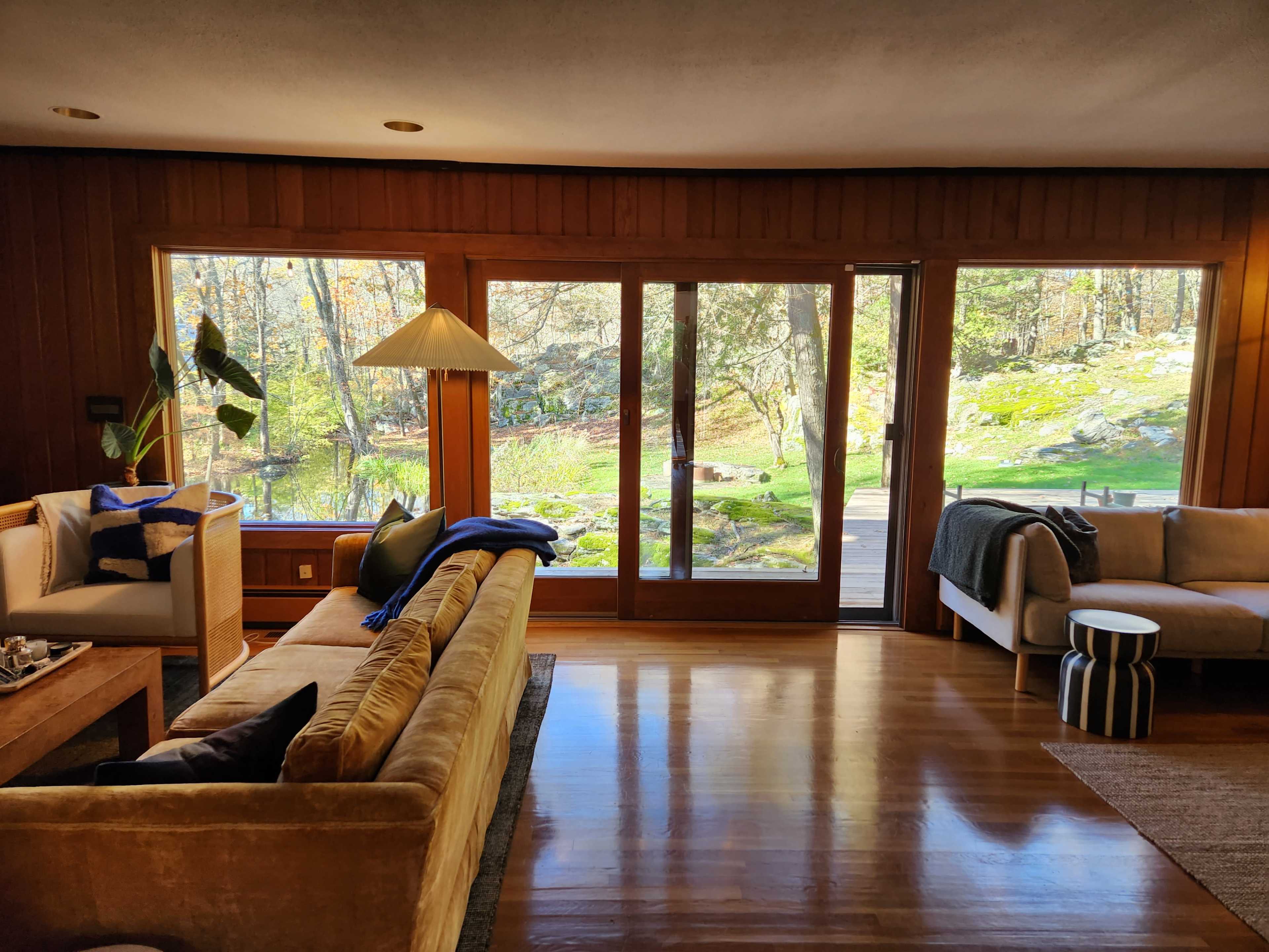 A spacious living room features large windows overlooking a natural landscape, with a mix of comfortable seating and decorative elements.