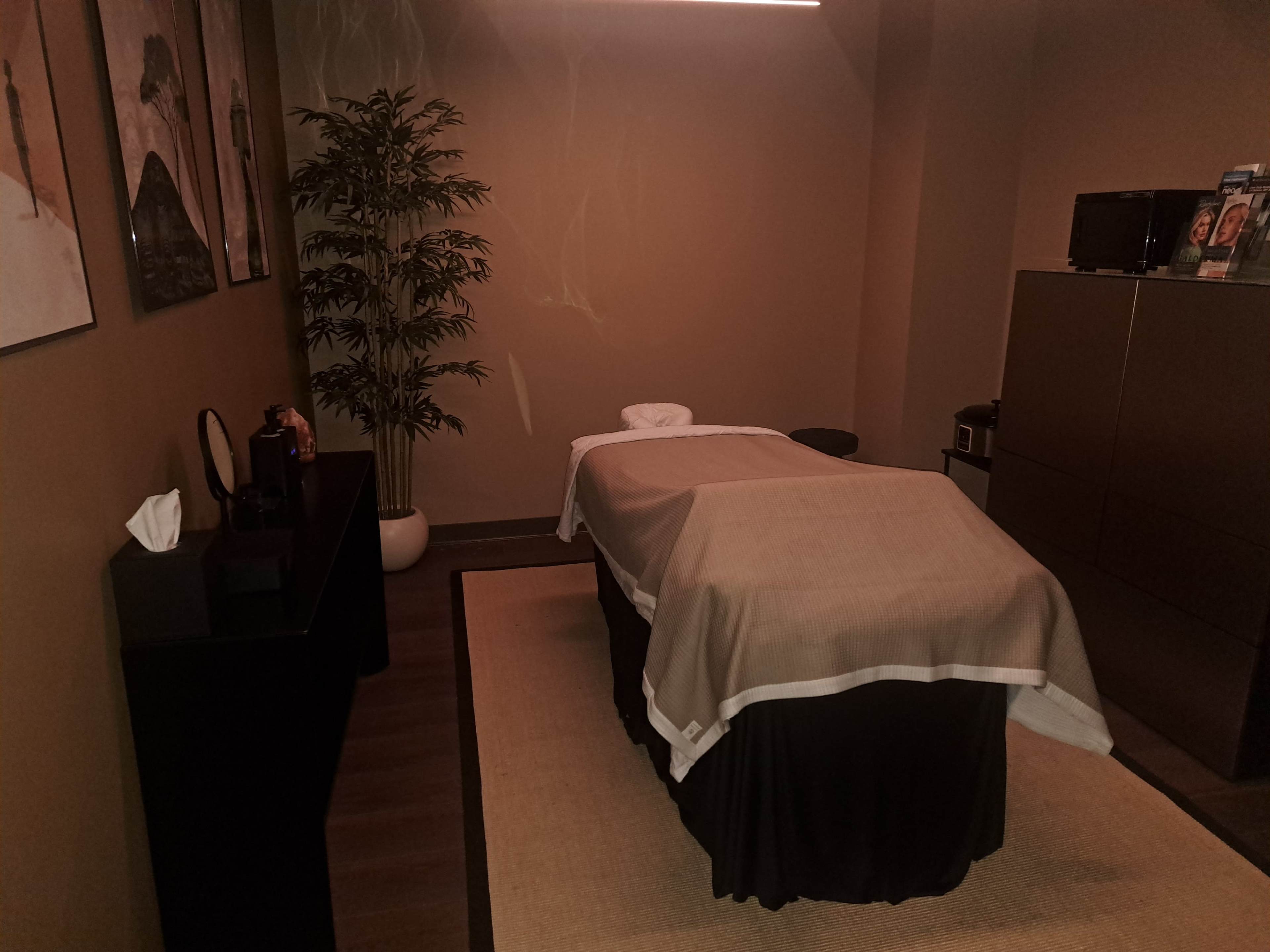 The image shows a massage room with a massage table covered by a sheet, a potted plant, and dark-colored furniture against a brown wall.