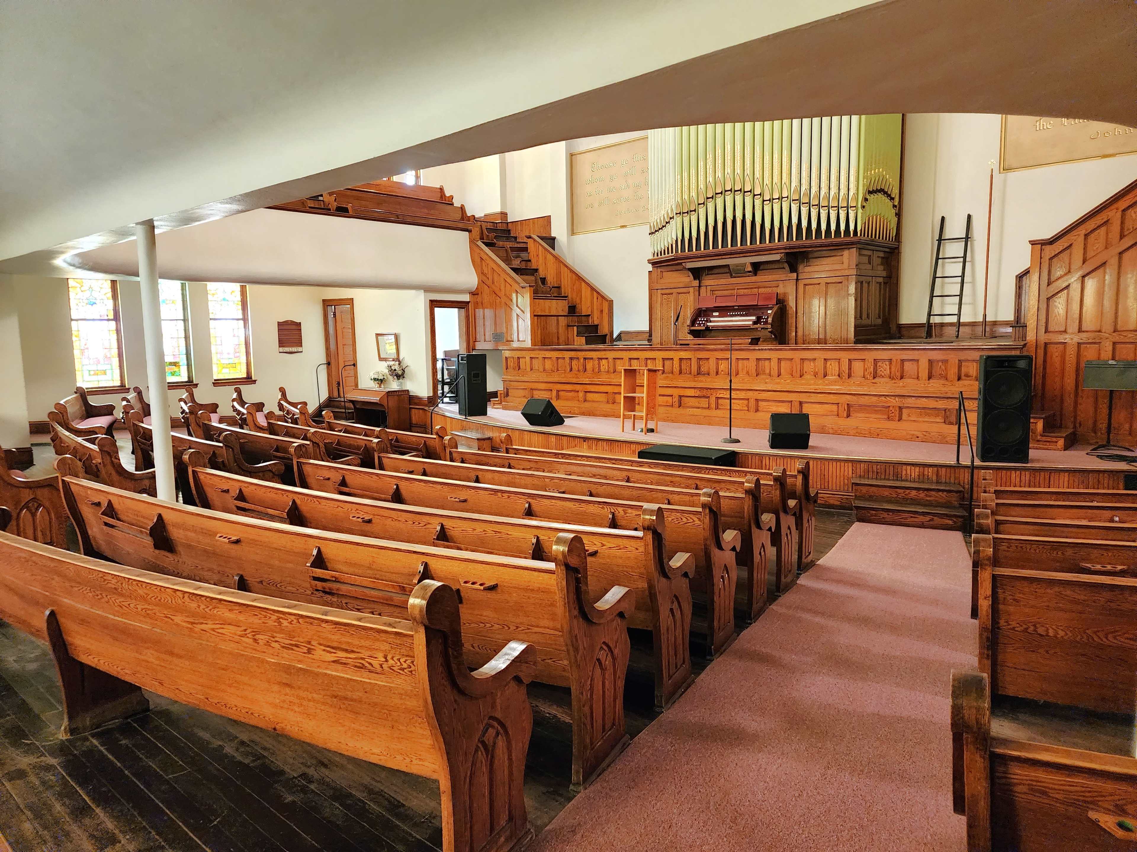 A wooden-paneled church interior with a row of pews facing a stage and an organ at the front.