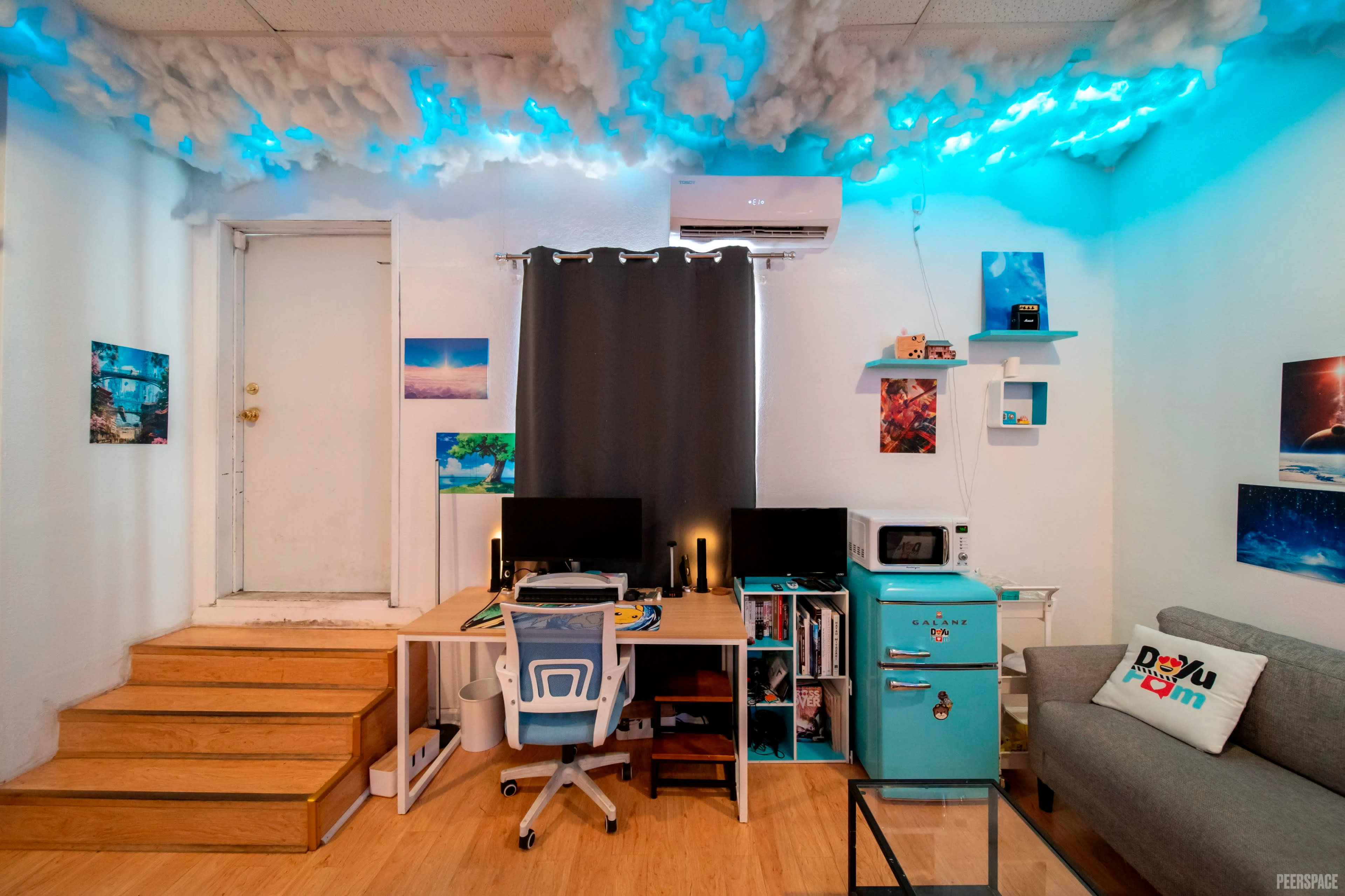 The image shows a brightly colored room with cloud-themed decor on the ceiling, a desk with two computer monitors, an old-style refrigerator, and a couch.