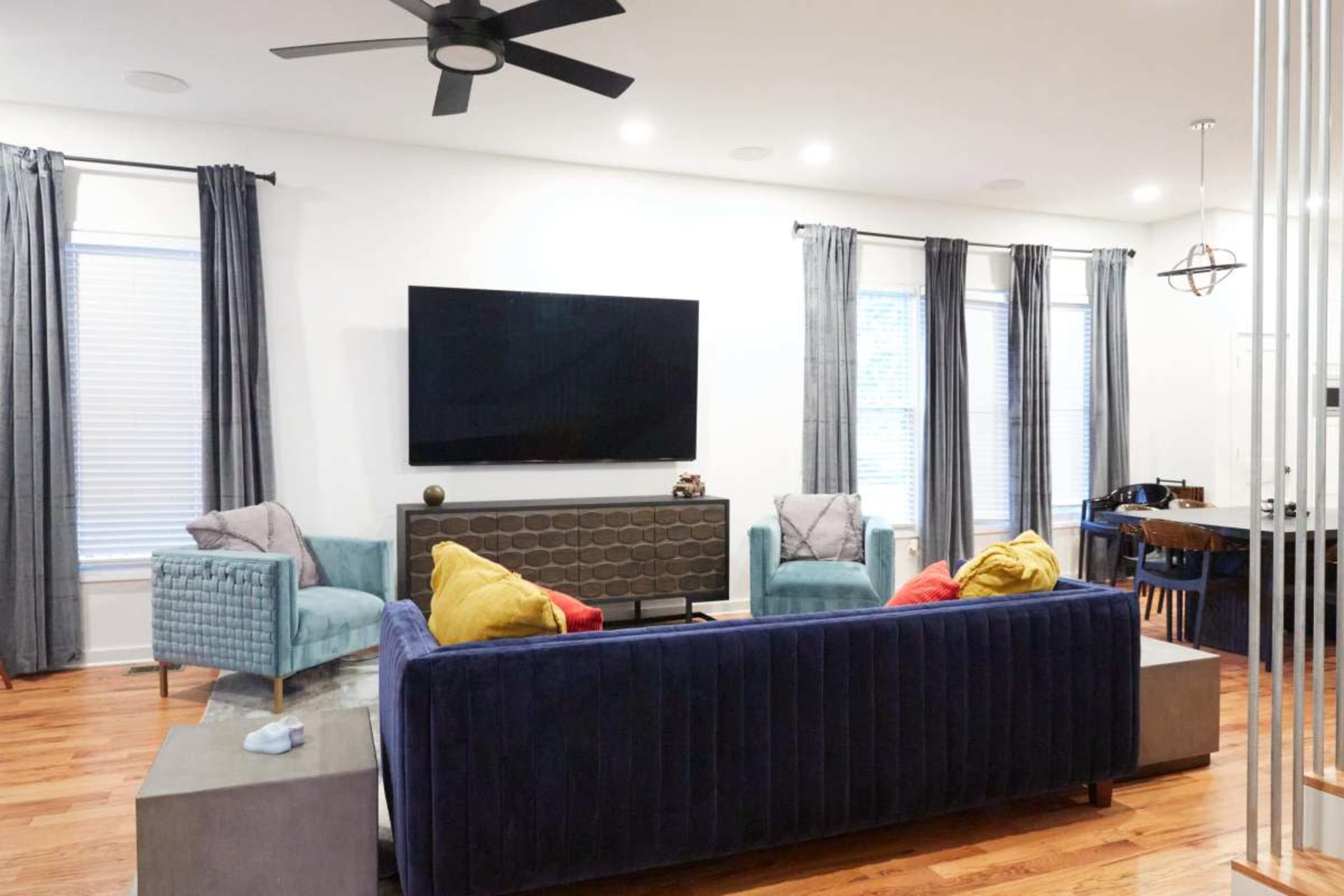 The image shows a modern living room featuring a dark blue sectional sofa, two light blue chairs, a large flat-screen TV mounted on the wall, and a dining area with a table in the background.