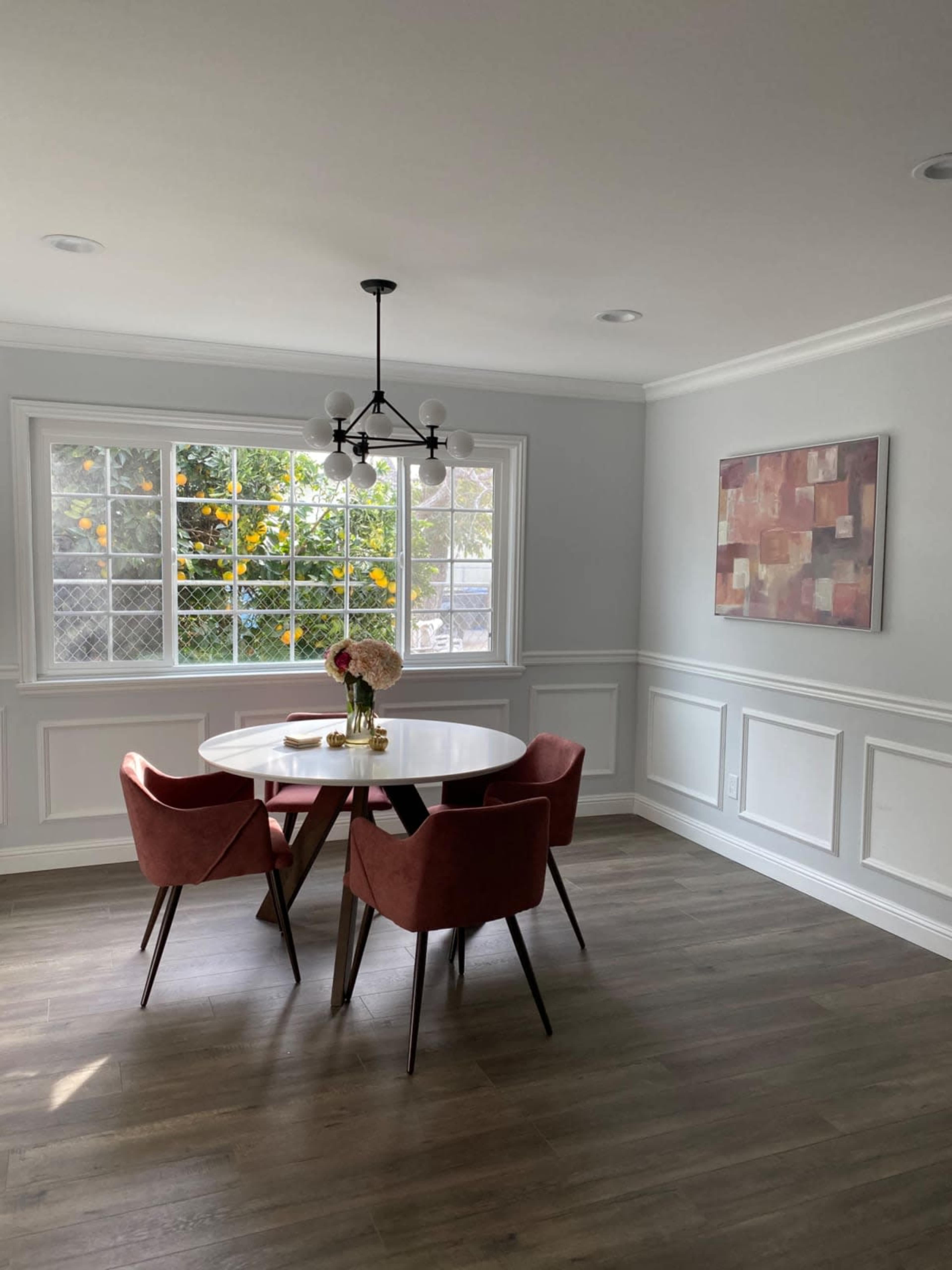 A modern dining area features a round white table surrounded by four pink chairs, next to a large window with greenery visible outside.