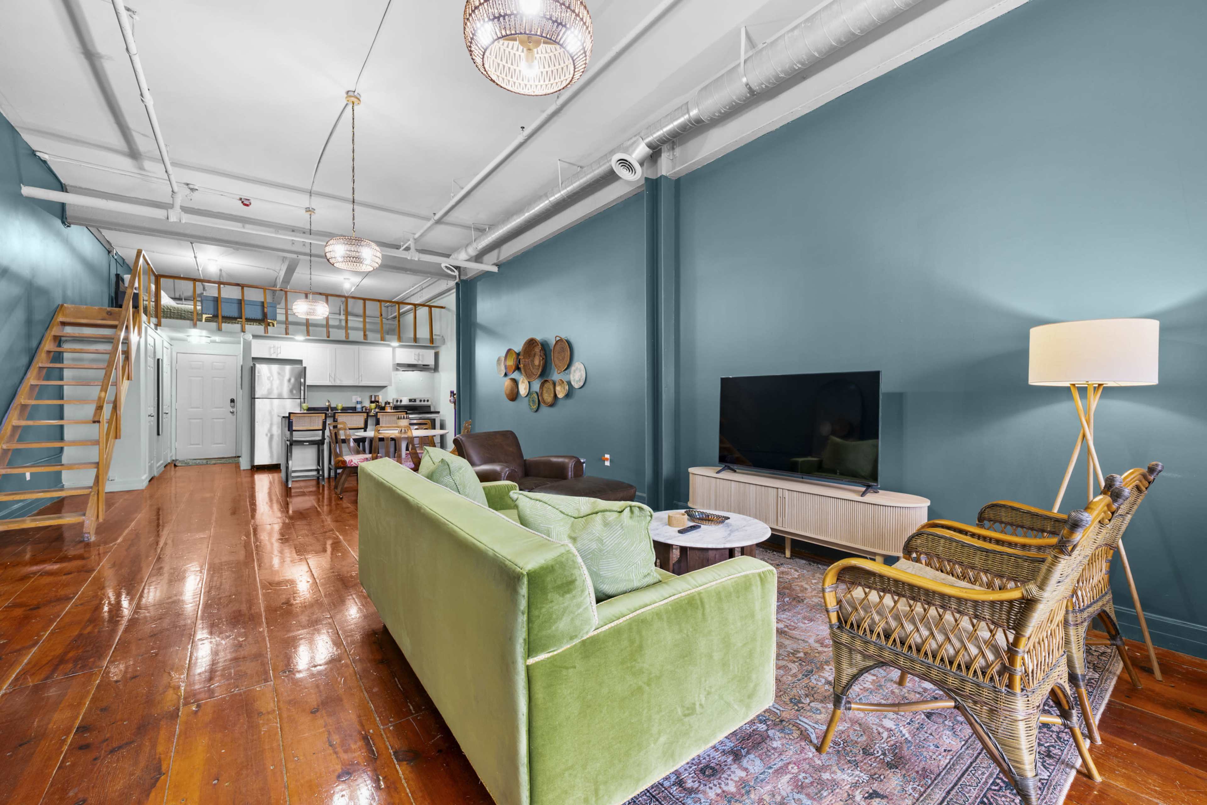 The image shows a spacious living area with a green sofa, two armchairs, a large television, and a dining area featuring a wooden table and chairs, set against a teal wall.