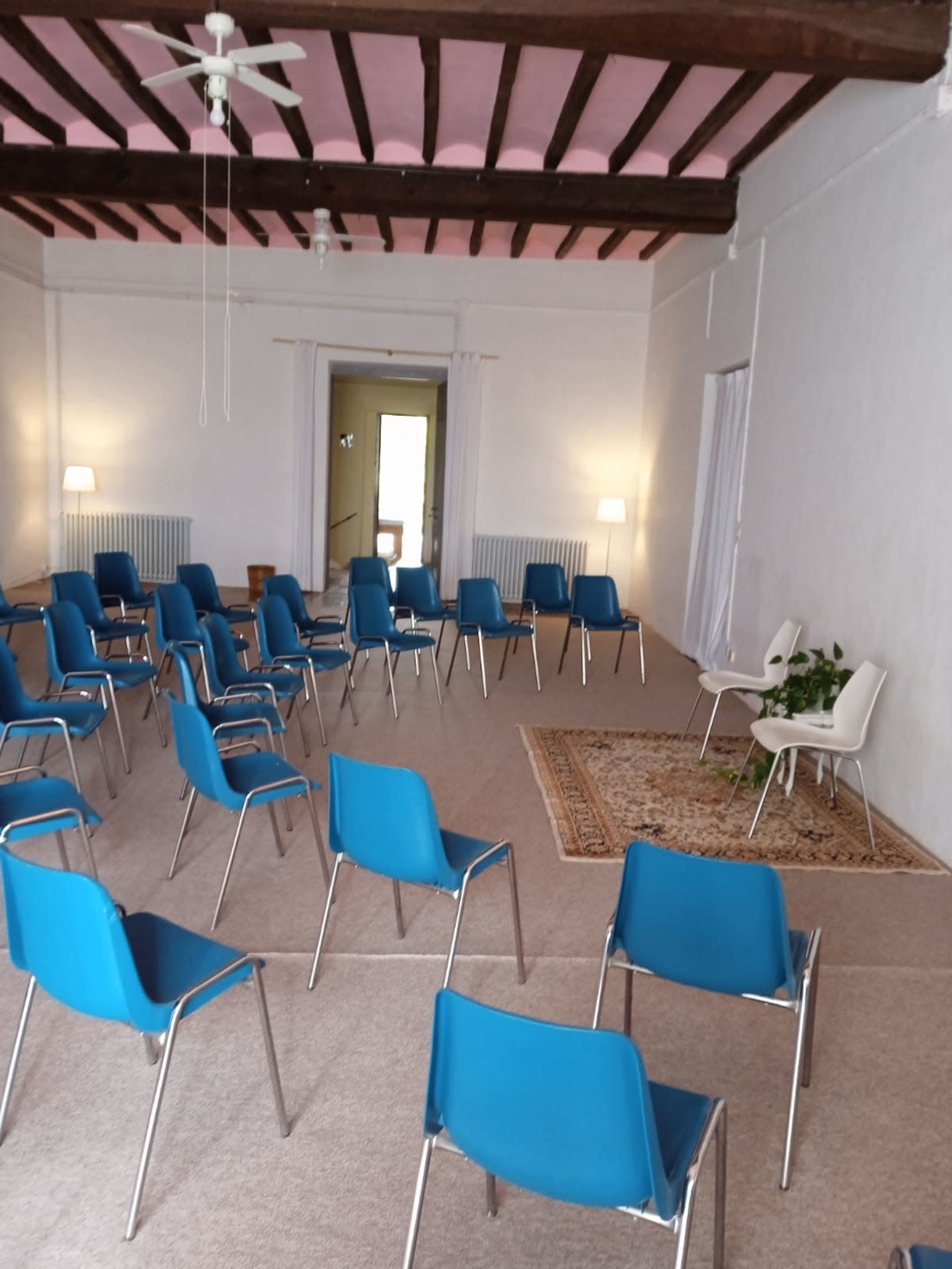A room with a carpeted floor features several blue chairs arranged in a circular formation around a small rug, with lamps and a doorway visible in the background.