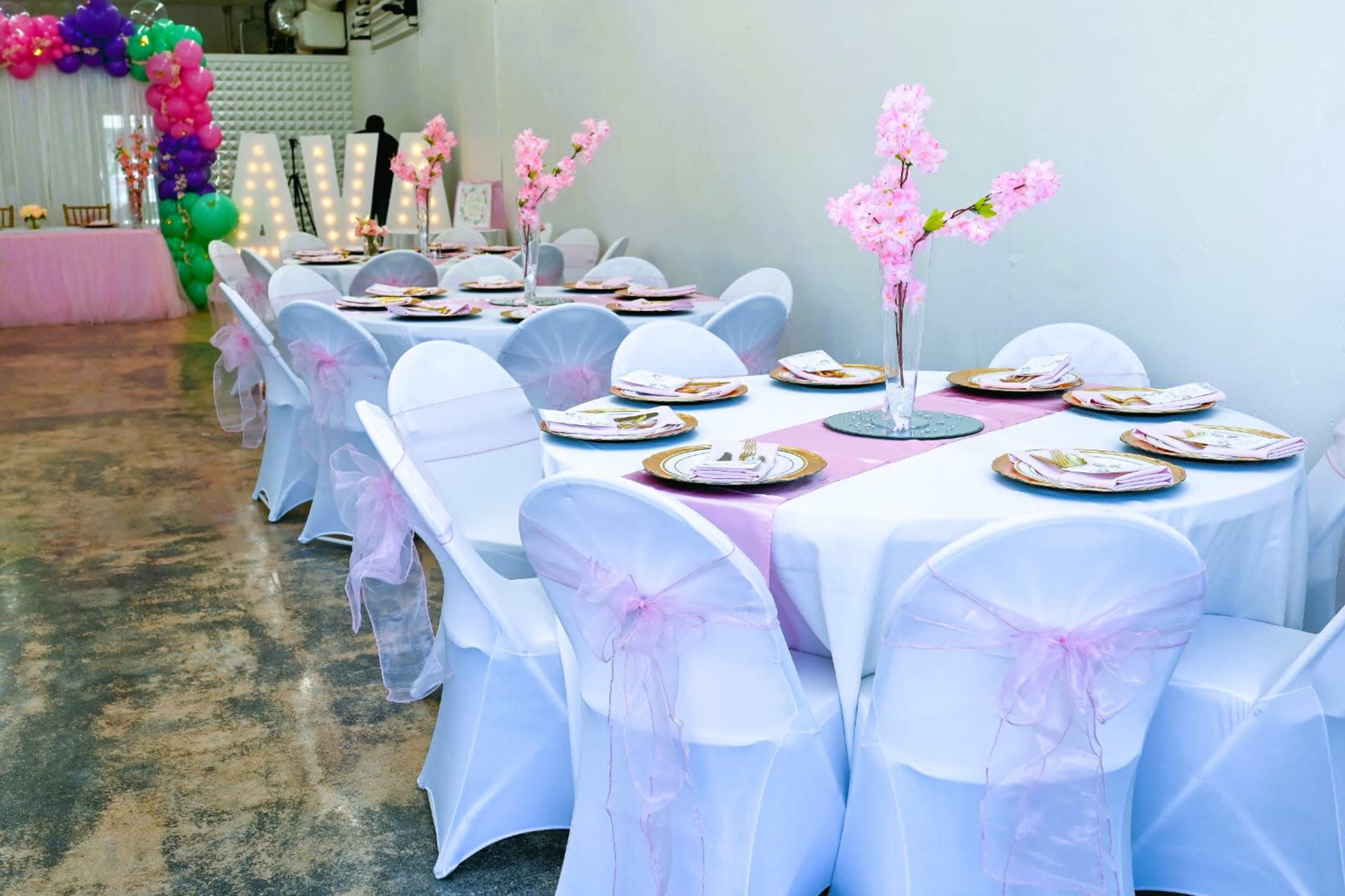 A banquet hall is set up with white tablecloths and chairs adorned with pink bows, featuring floral centerpieces on each table.