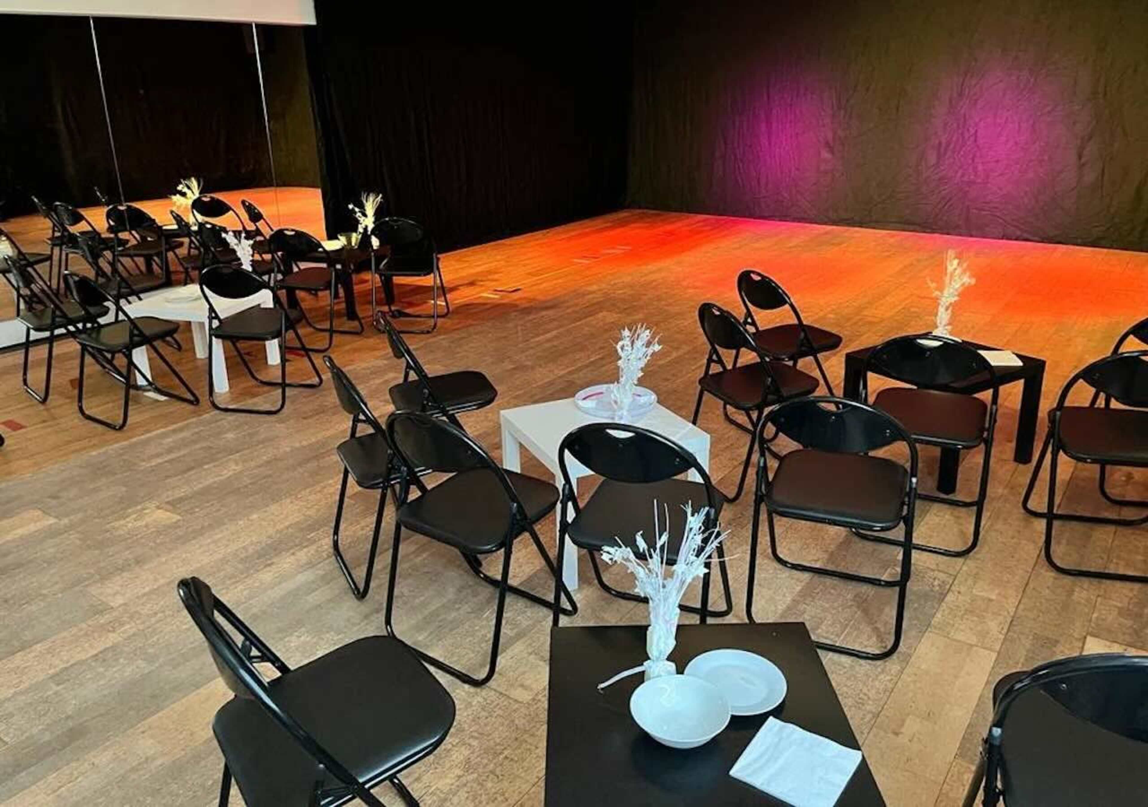 The image shows a stage setup with black folding chairs arranged in a semi-circle around a small white table, featuring minimalist centerpieces, in a dimly lit space.