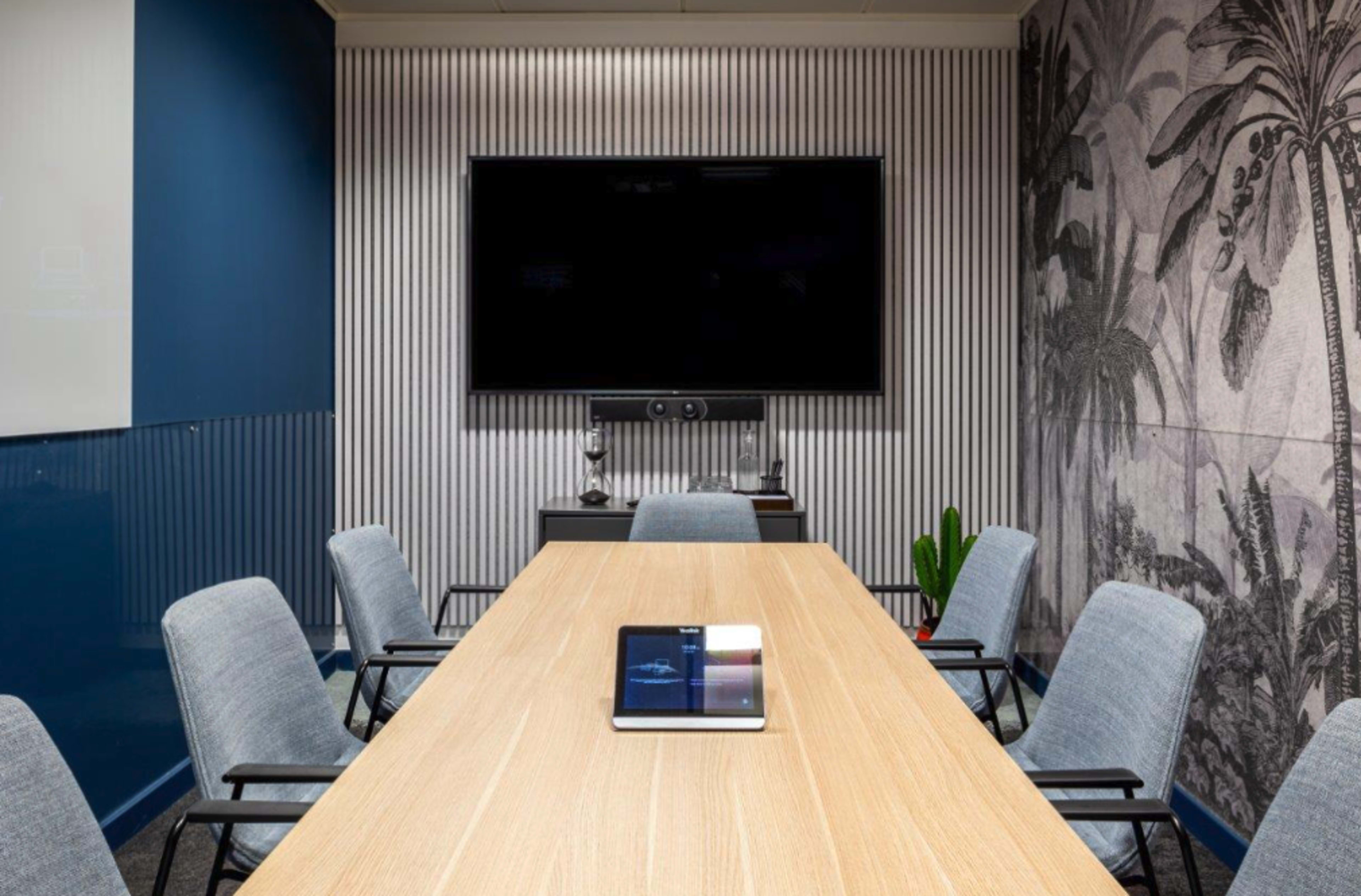 A conference room features a long wooden table, several gray chairs, a large wall-mounted TV, and a tablet on the table.