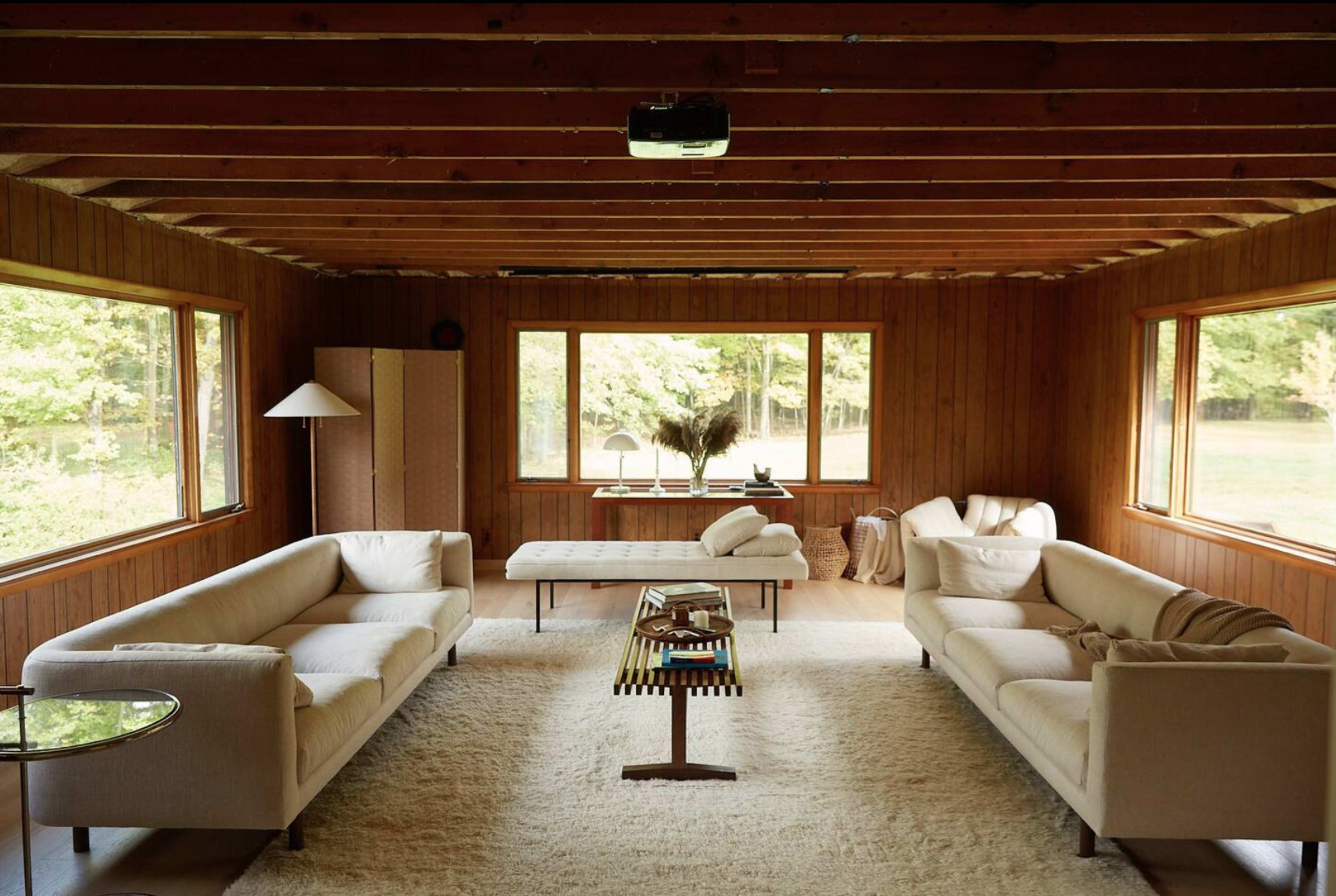 A cozy living room features two beige sofas facing a coffee table, with large windows overlooking a green landscape and wooden walls.