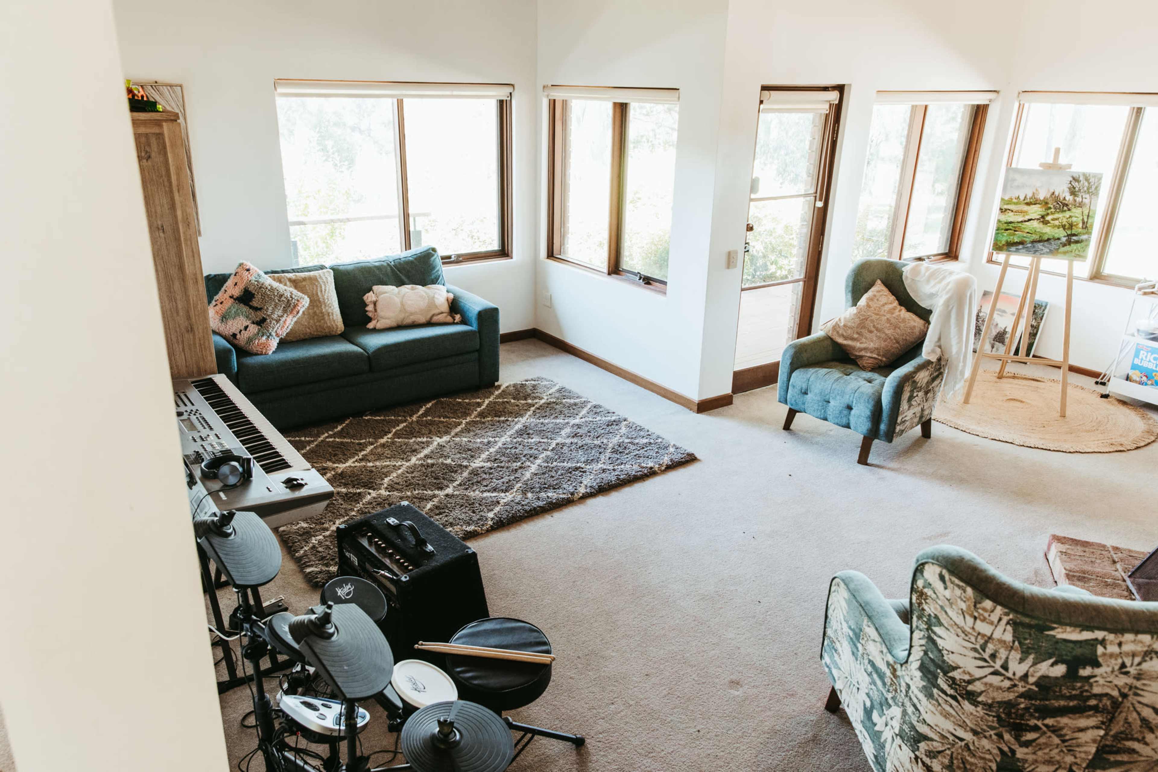 A spacious living room with a green couch, an armchair, a keyboard, and large windows allowing natural light to enter.