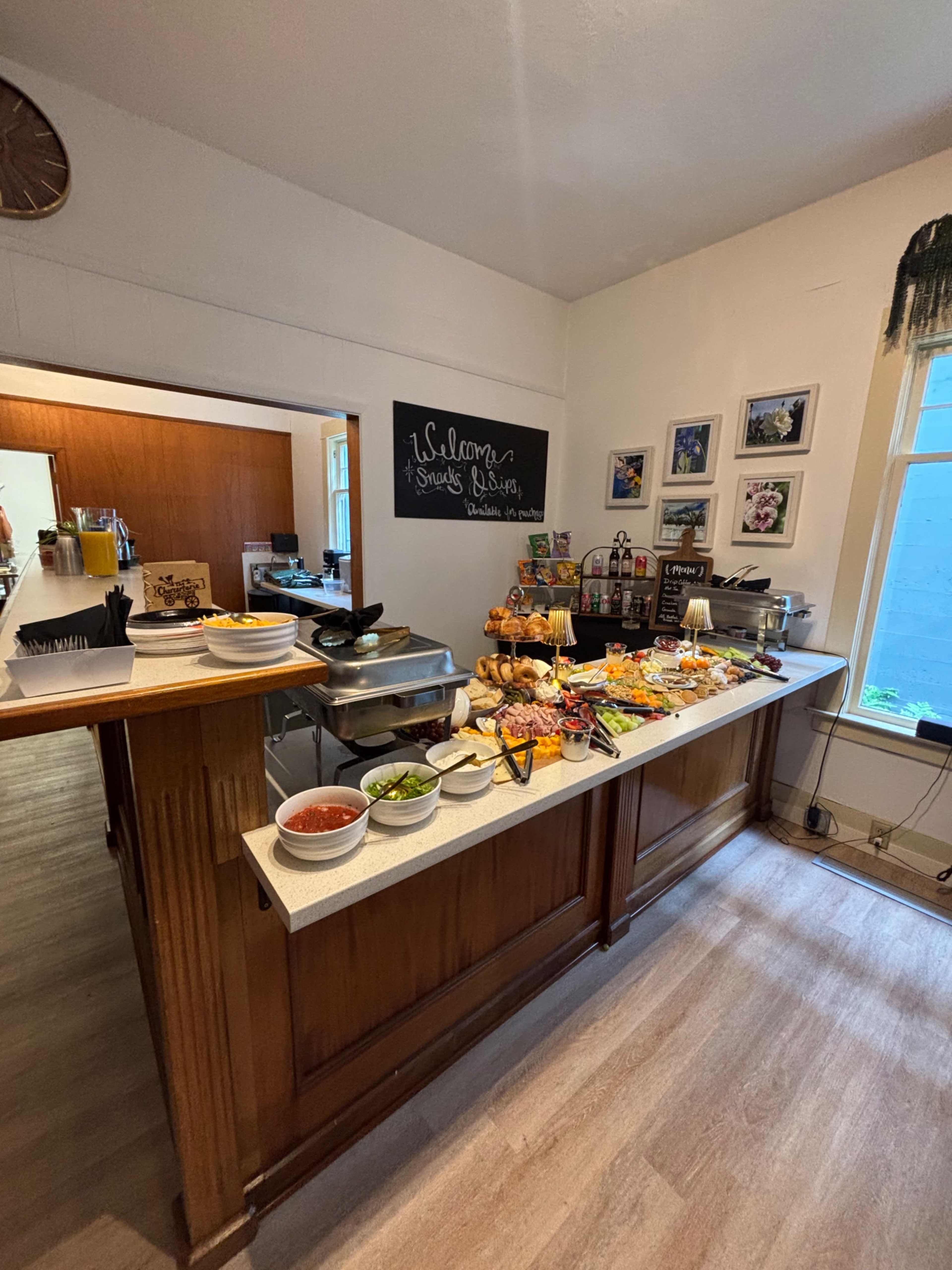 A buffet table is set up with various dishes, including fruits, vegetables, and condiments, against a wall adorned with framed pictures and a welcoming message.