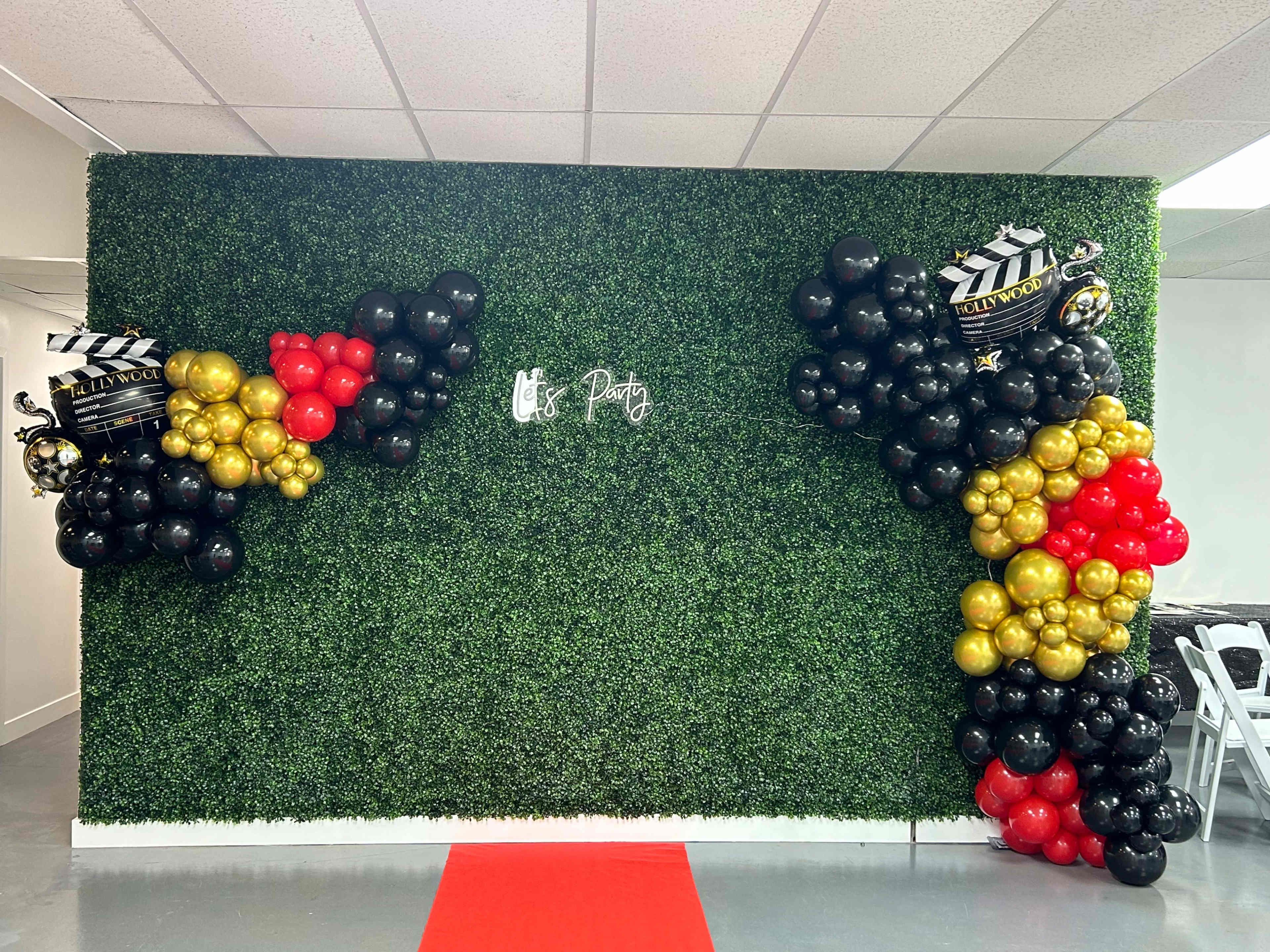 The scene features a party backdrop made of artificial greenery, adorned with balloon arrangements in black, gold, and red, and a red carpet leading up to the display.