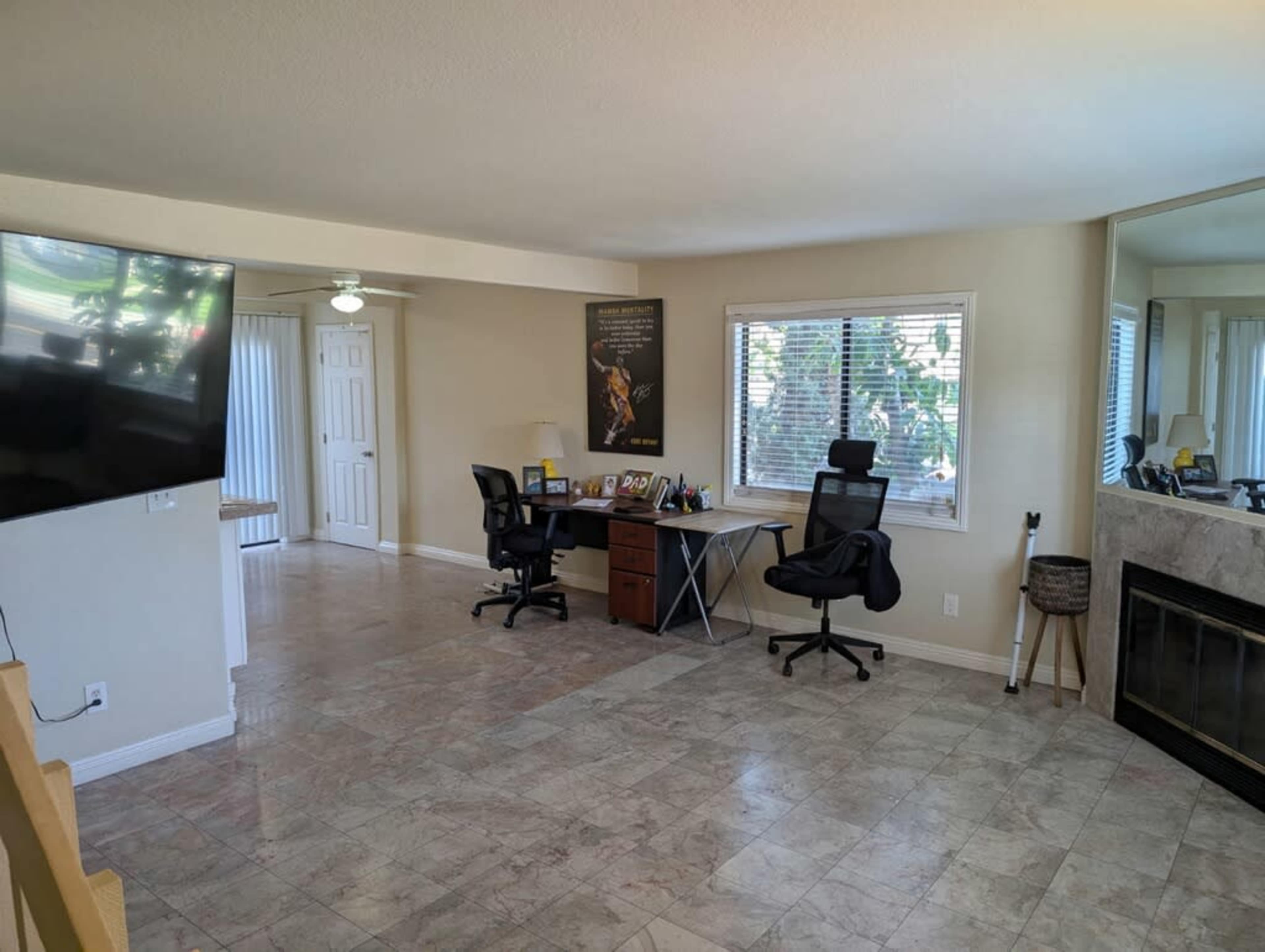 The image shows a spacious living area featuring a desk with two chairs, a television mounted on the wall, and a fireplace with a mirror adjacent to it.