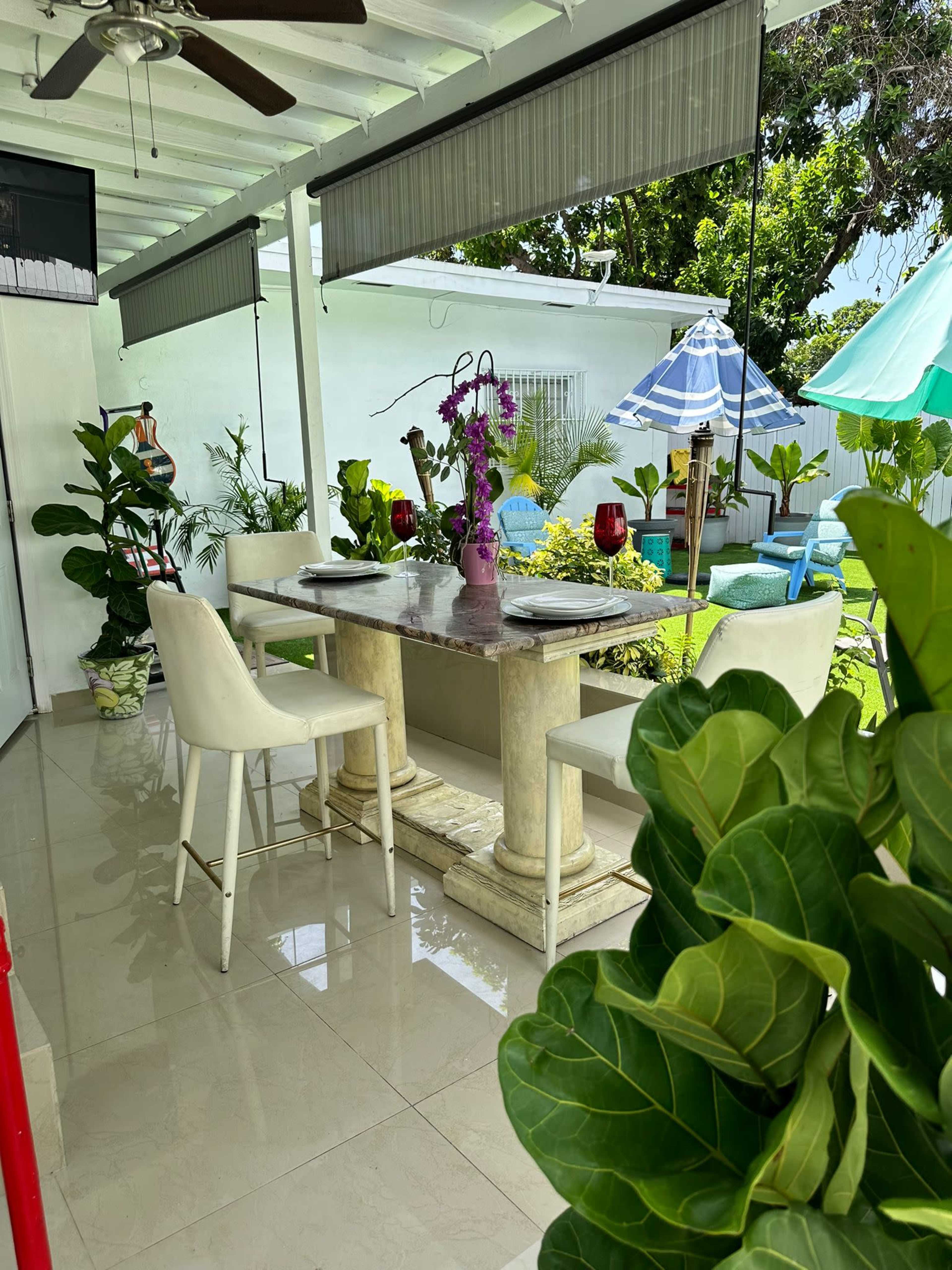 A patio area features a table with chairs, surrounded by potted plants and patio furniture under a covered roof.