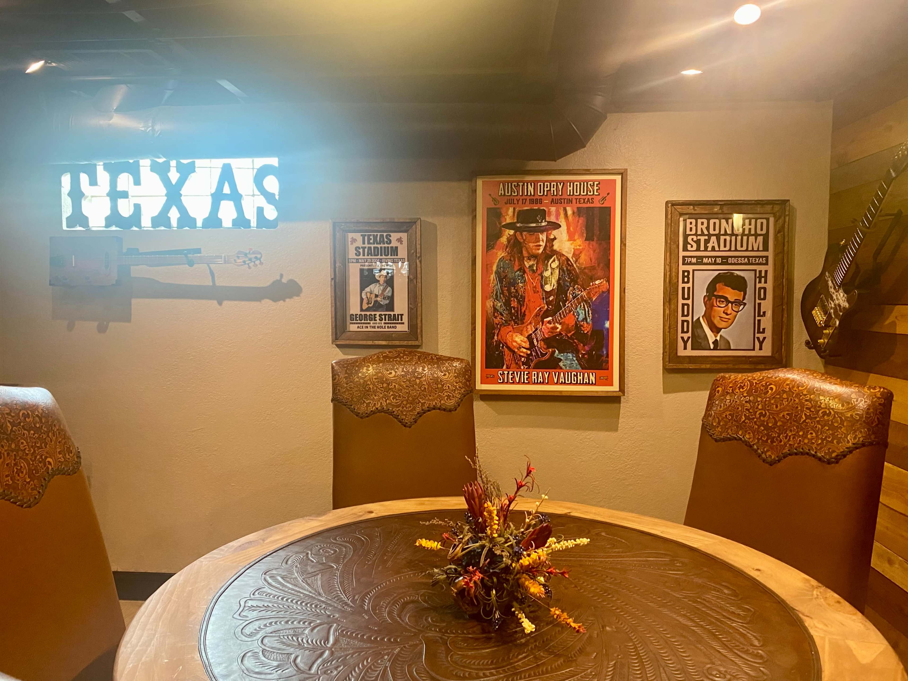 A rustic dining area features a round table with a floral centerpiece, framed artwork of Texas music legends on the walls, and wooden chairs.