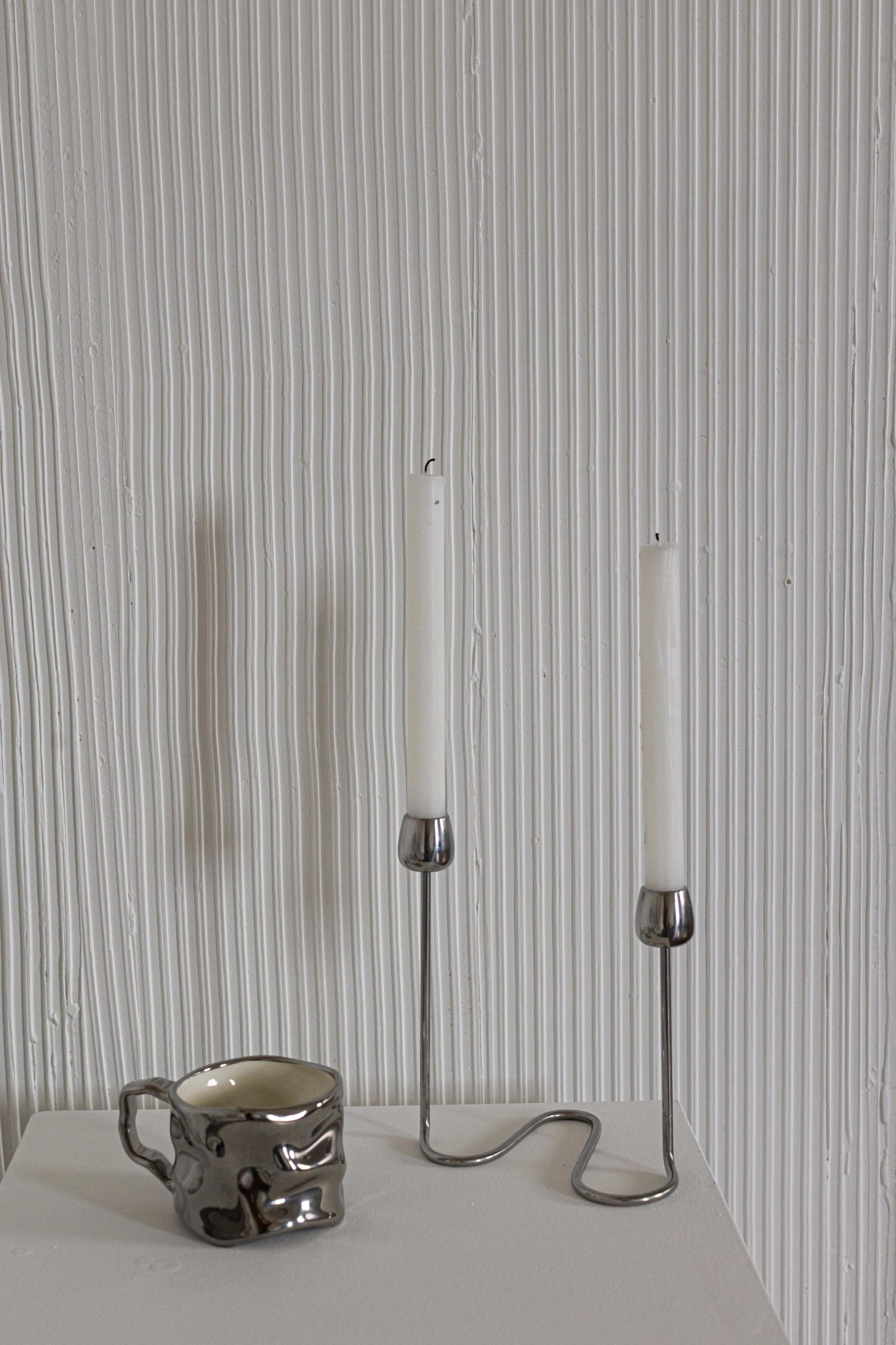 A metallic candle holder with two white candles stands next to a textured gray ceramic mug on a white table against a ridged white wall.