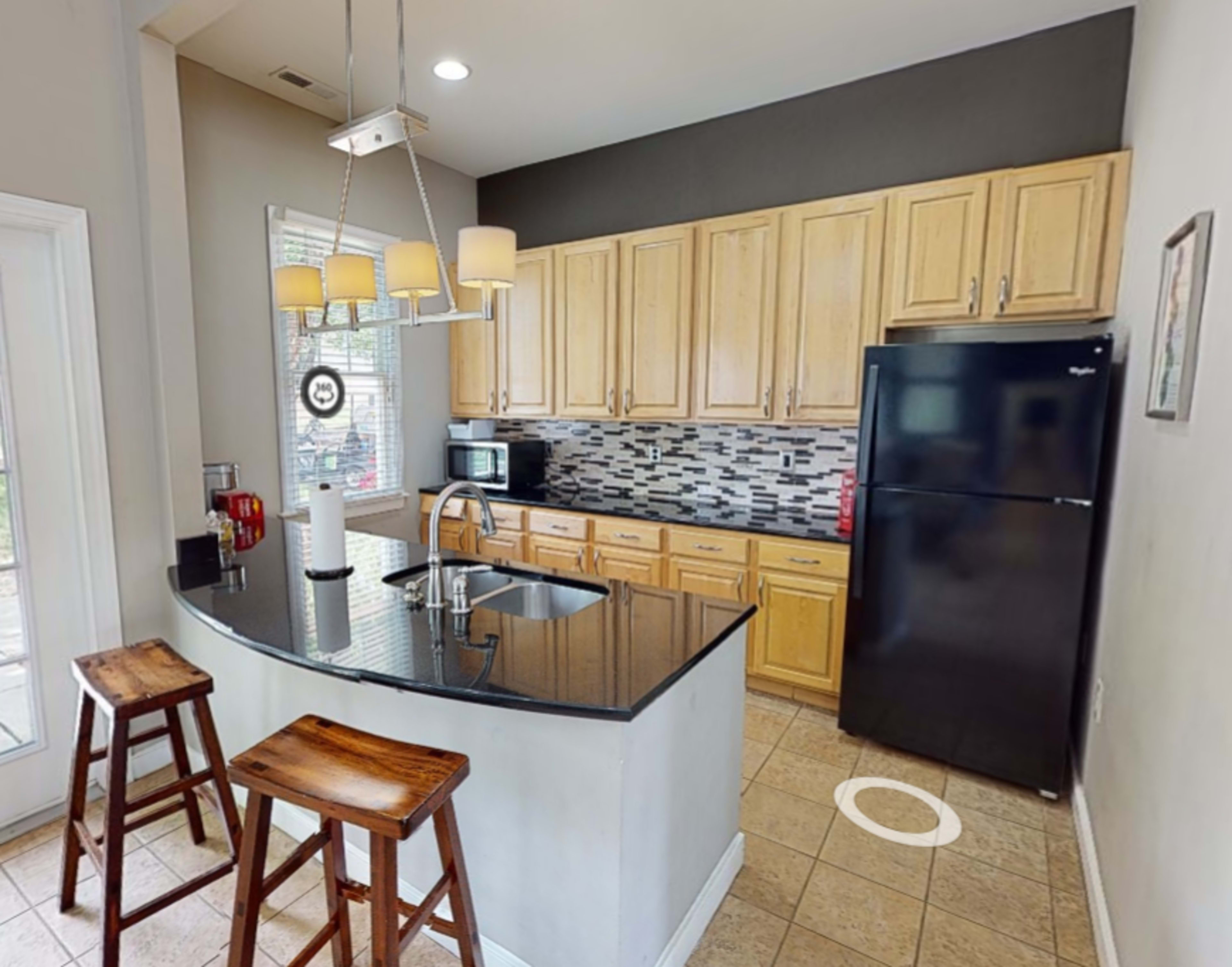 A modern kitchen featuring a curved granite countertop with bar stools, wooden cabinets, and a black refrigerator.