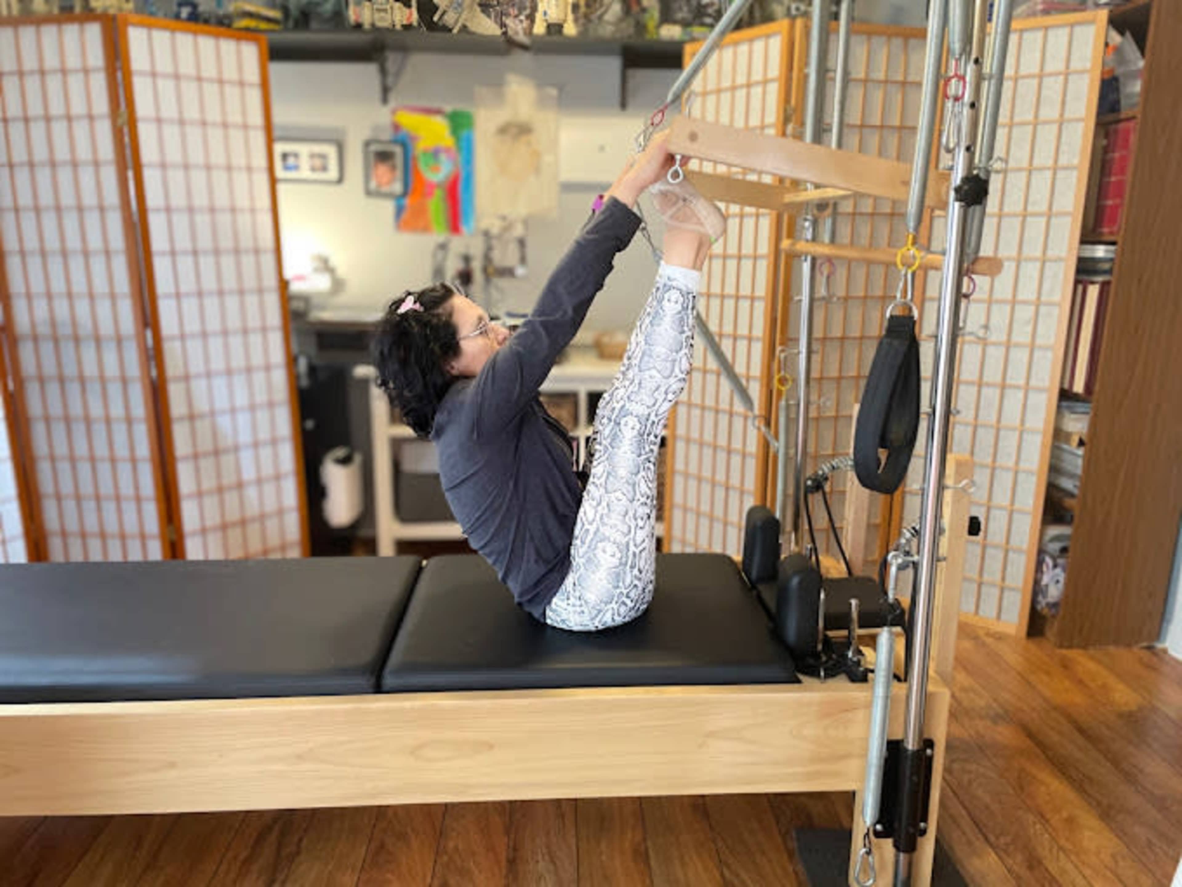 A person performs an exercise on a Pilates reformer in a studio setting with wooden floors and decorative screens in the background.