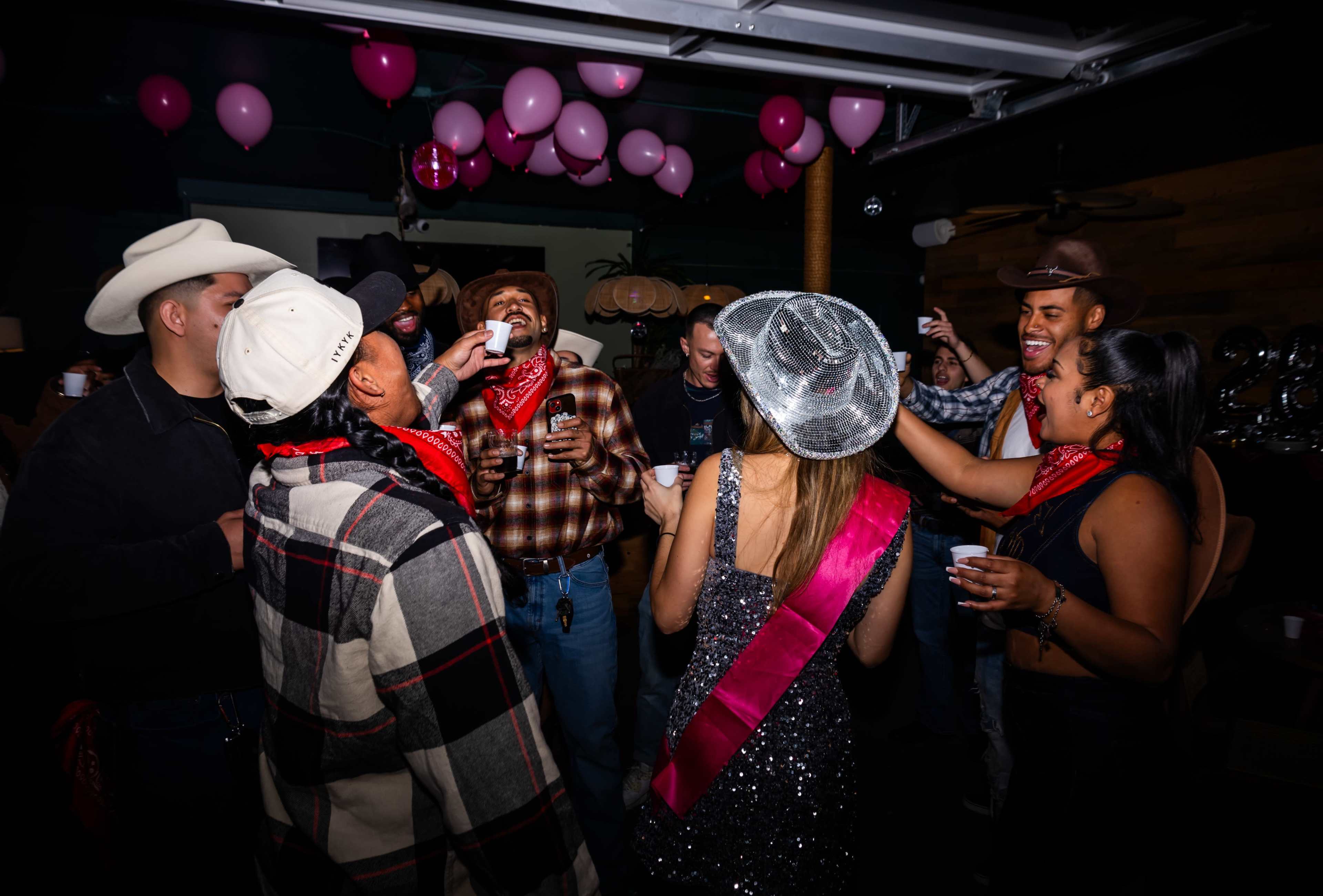 A group of people in cowboy attire celebrate at a party with balloons and drinks.