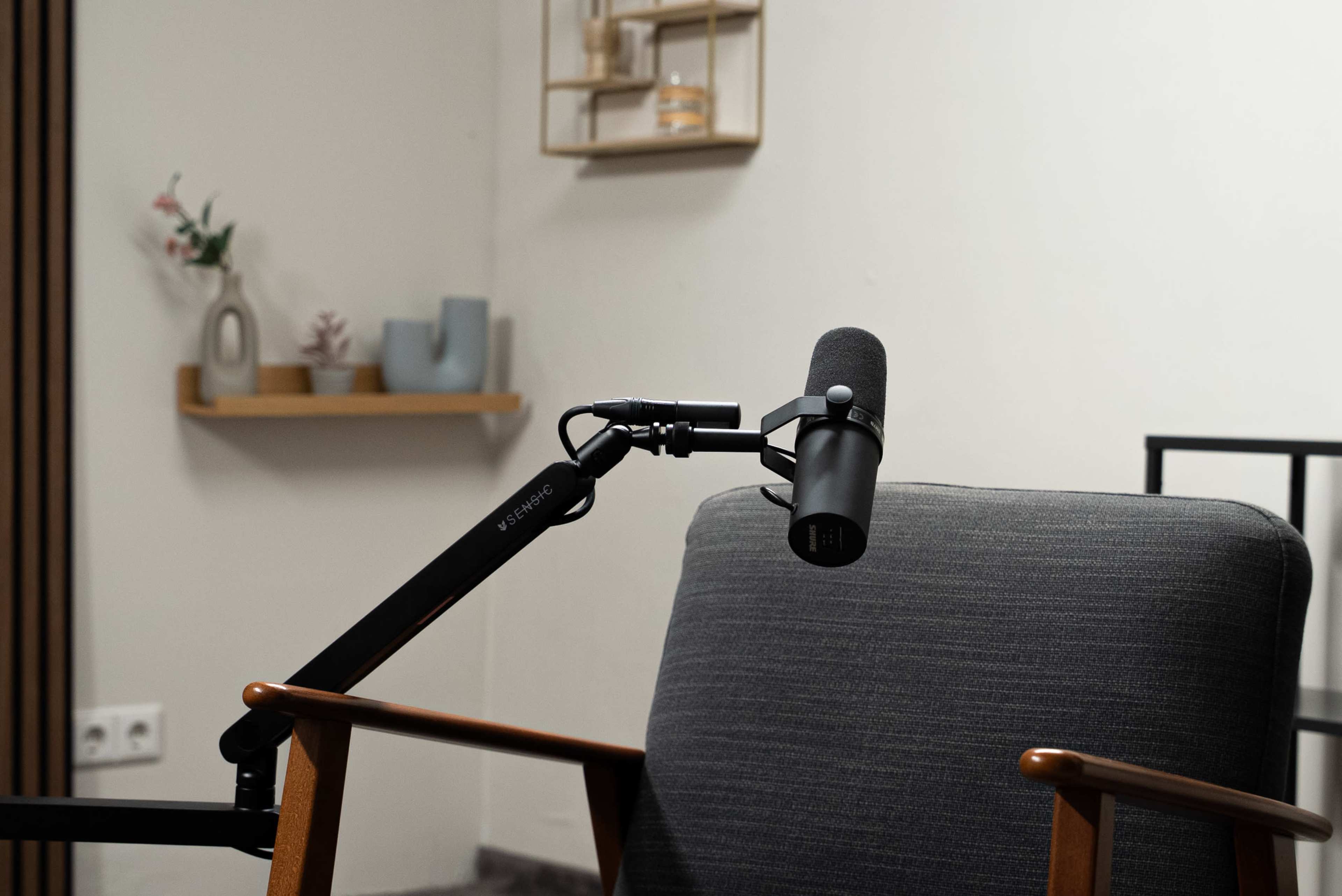 A microphone is mounted on an adjustable arm near a gray armchair in a minimalist room with decorative shelves.