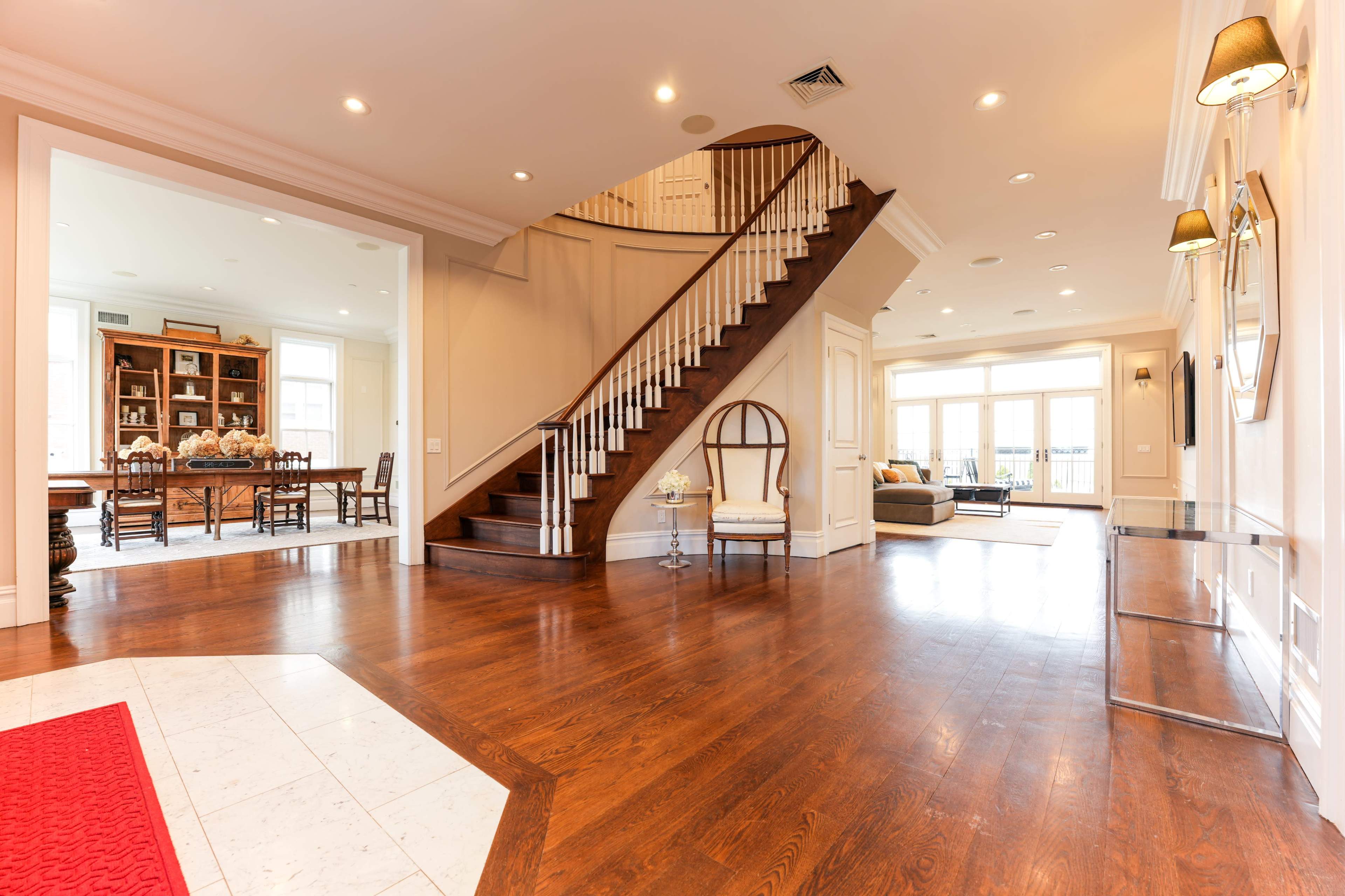 A spacious foyer with a wooden staircase leading to the upper level, adjoining a dining area and a living room with large windows.