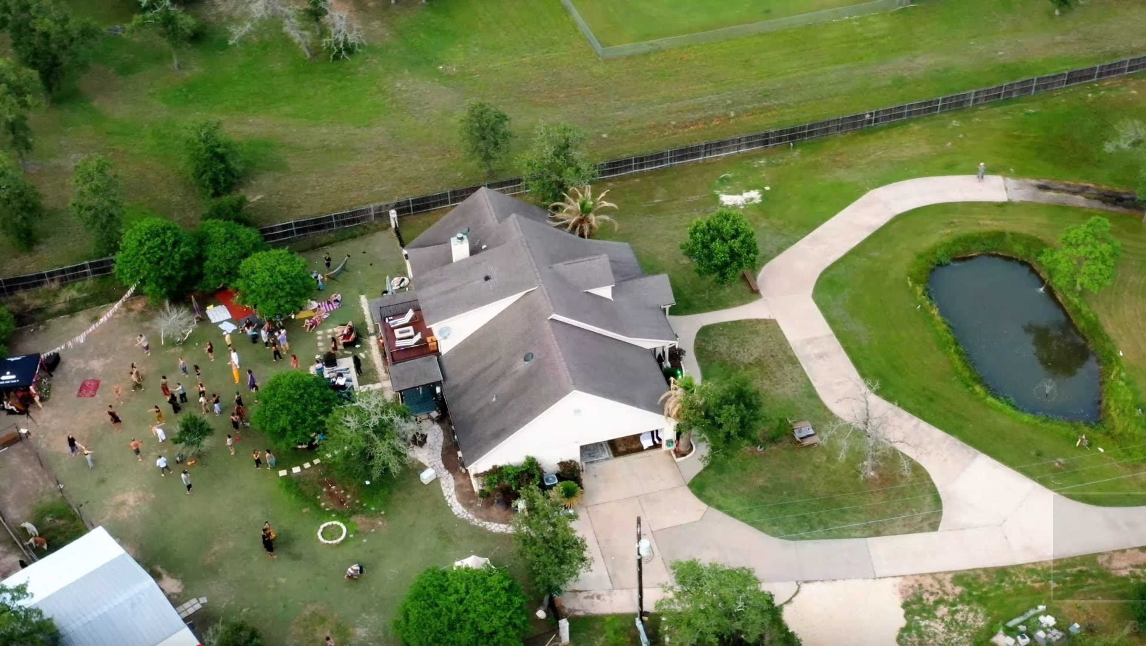 An aerial view of a house with a large outdoor area featuring a gathering of people, surrounded by trees and a pond.