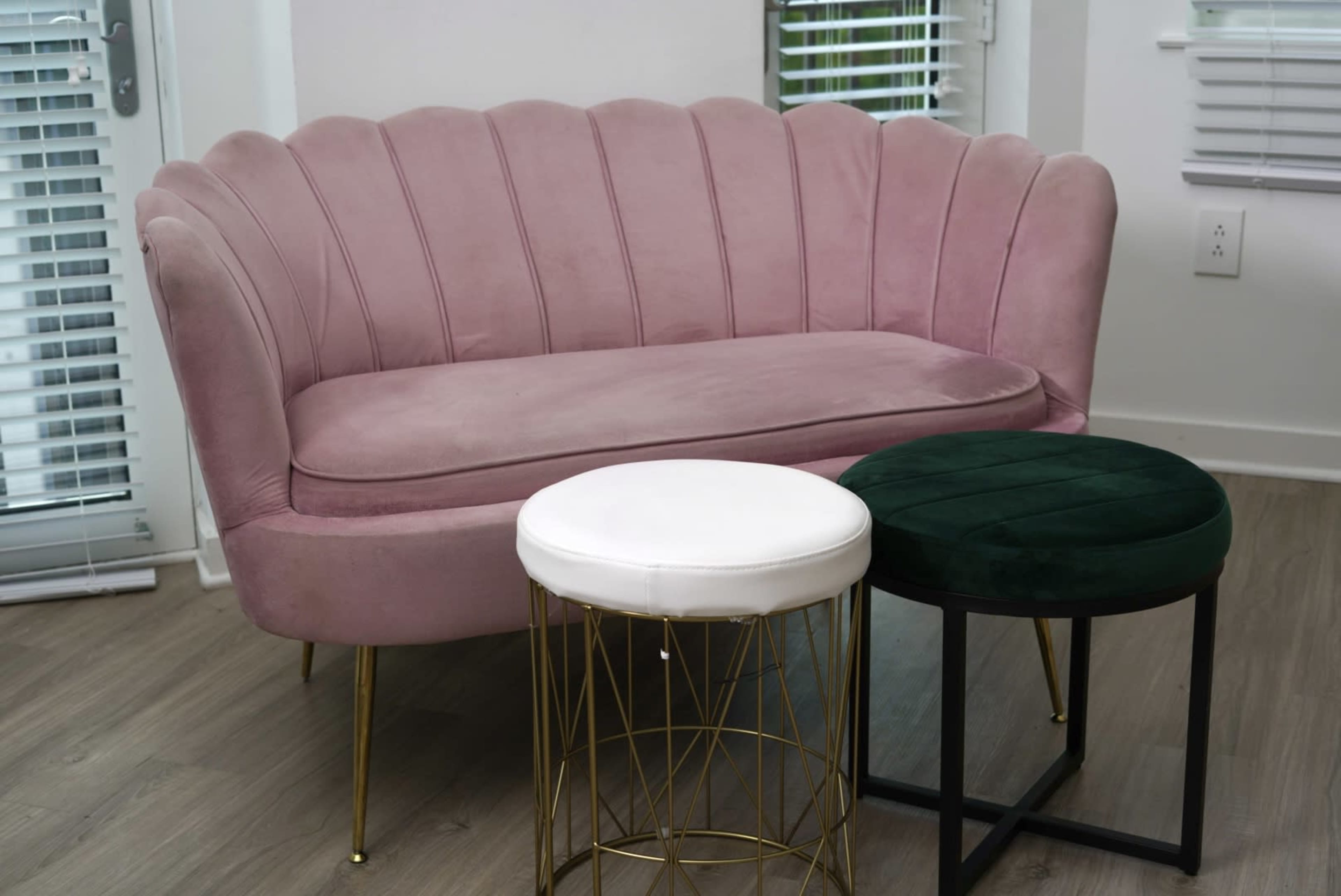 A pink velvet sofa with a scalloped backrest is positioned next to two round stools, one white and one green, with metallic and wooden bases.