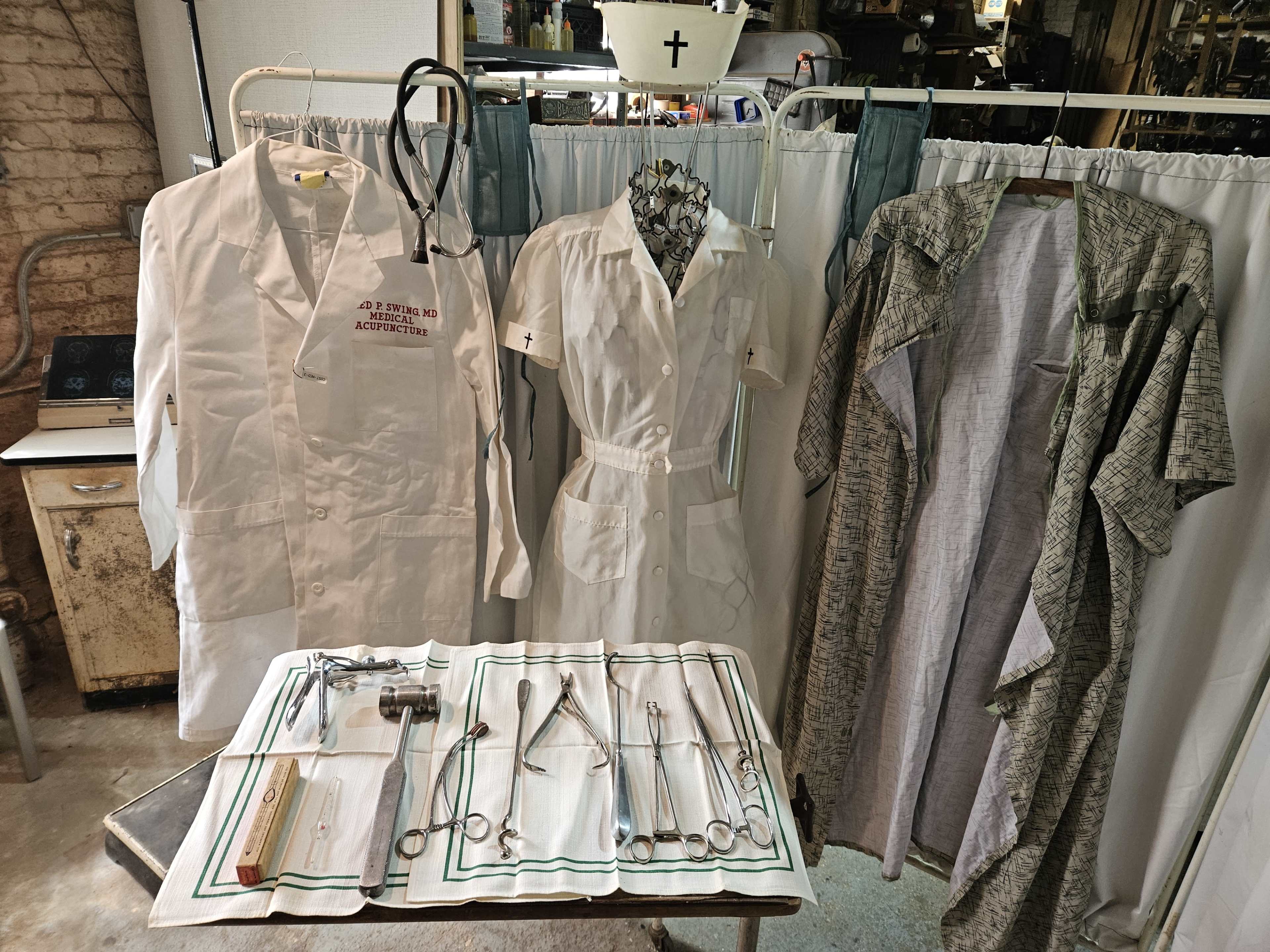 The image shows a table with various surgical instruments, surrounded by medical uniforms and a stethoscope displayed on a backdrop of draped fabric.
