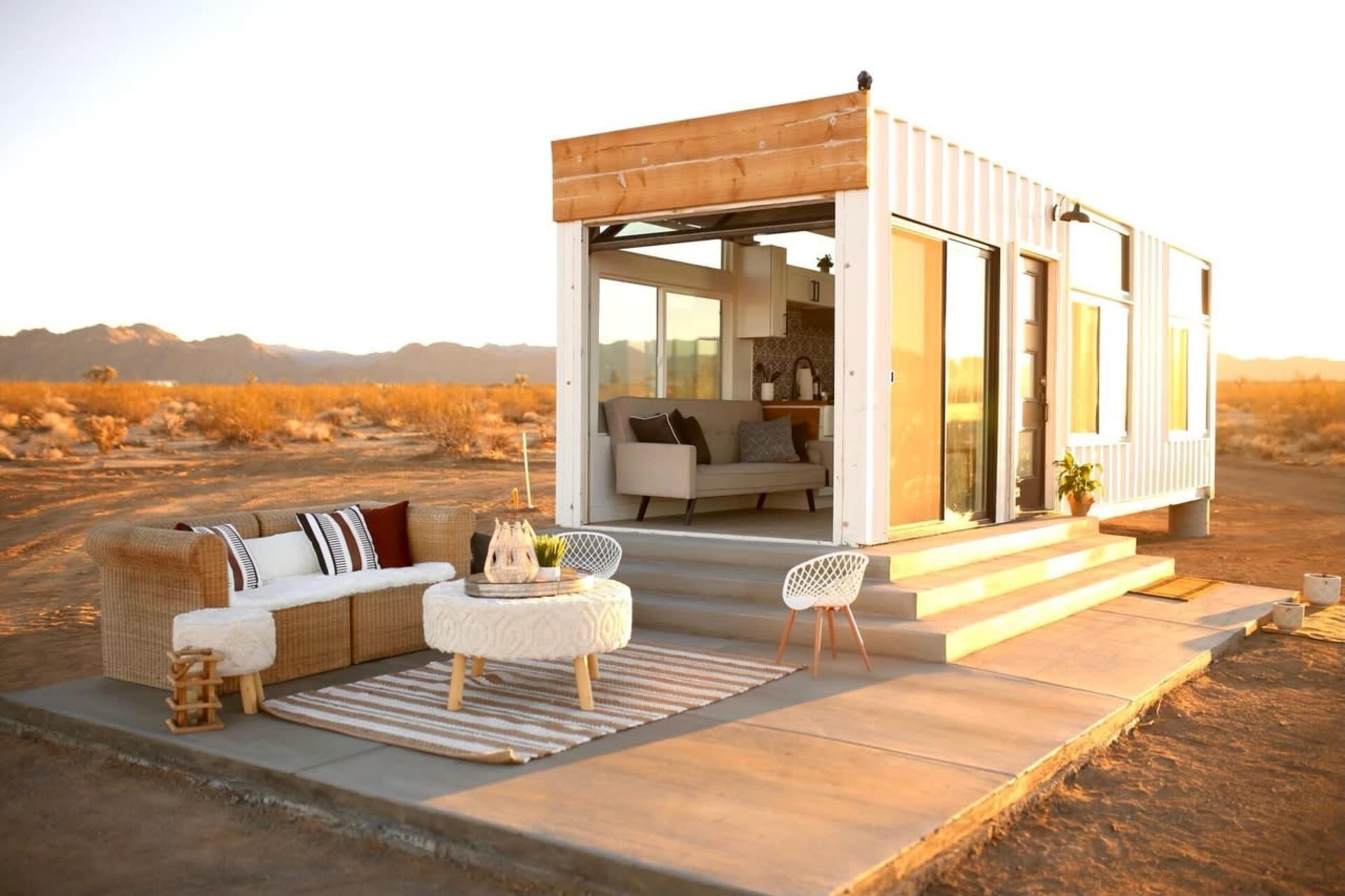 A modern container home is set on a concrete platform in a desert landscape, featuring large sliding glass doors and a simple outdoor seating area.