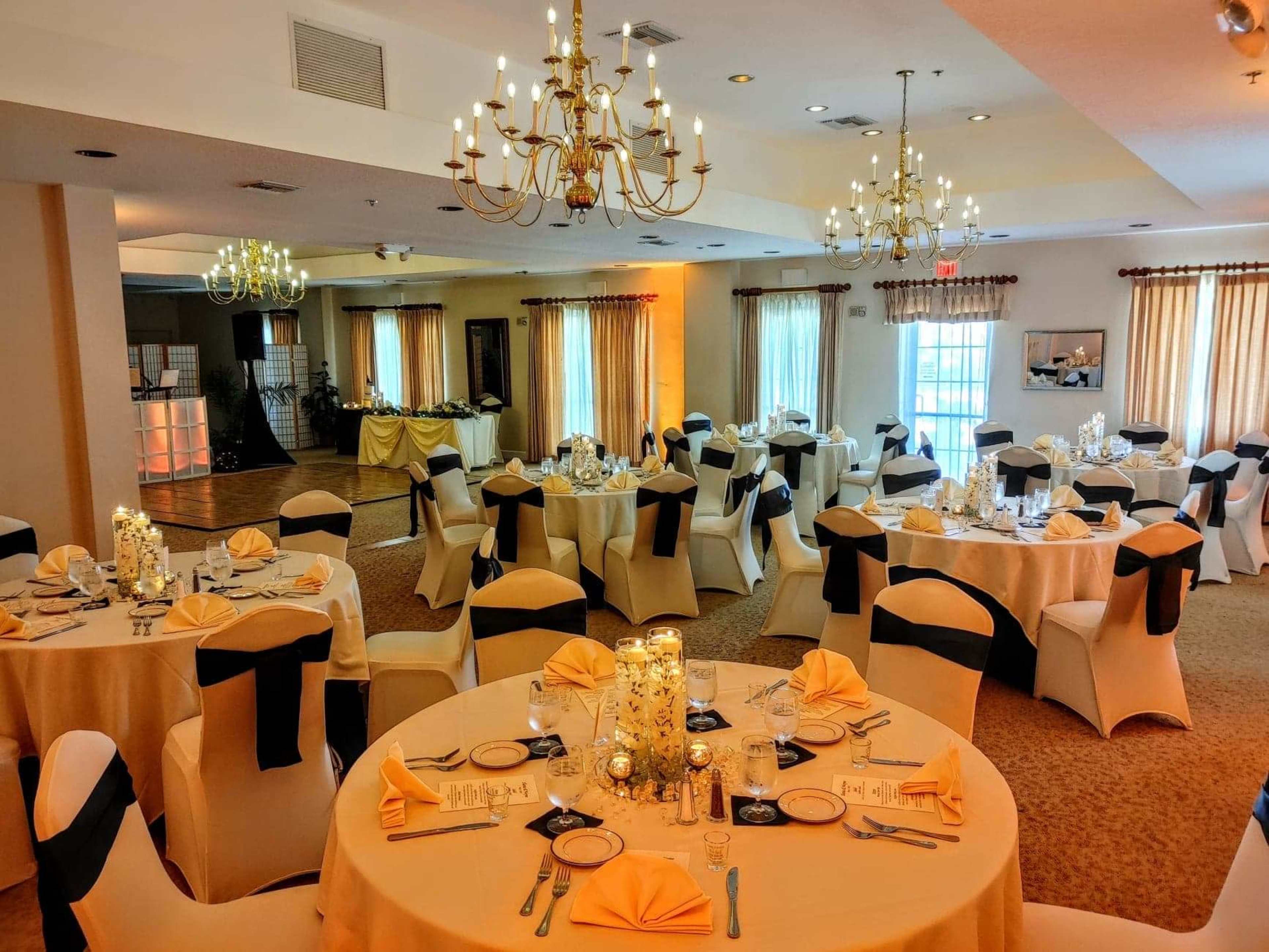 A banquet hall set up for dining, featuring round tables with beige tablecloths and black chair covers, surrounded by elegant chandeliers and decorated with floral arrangements and candles.